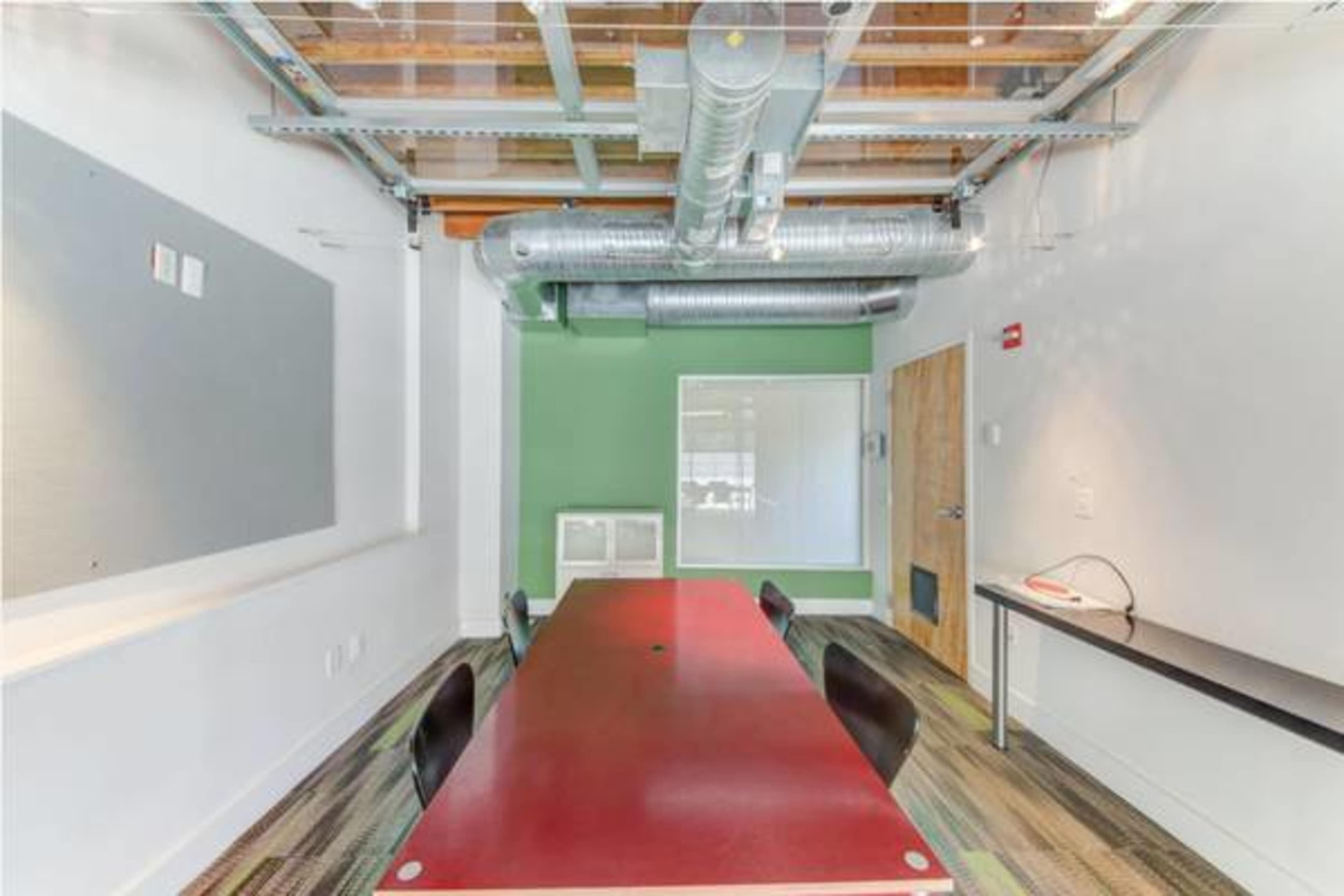The image shows a modern conference room featuring a long red table surrounded by black chairs, with green and grey walls and exposed ductwork above.
