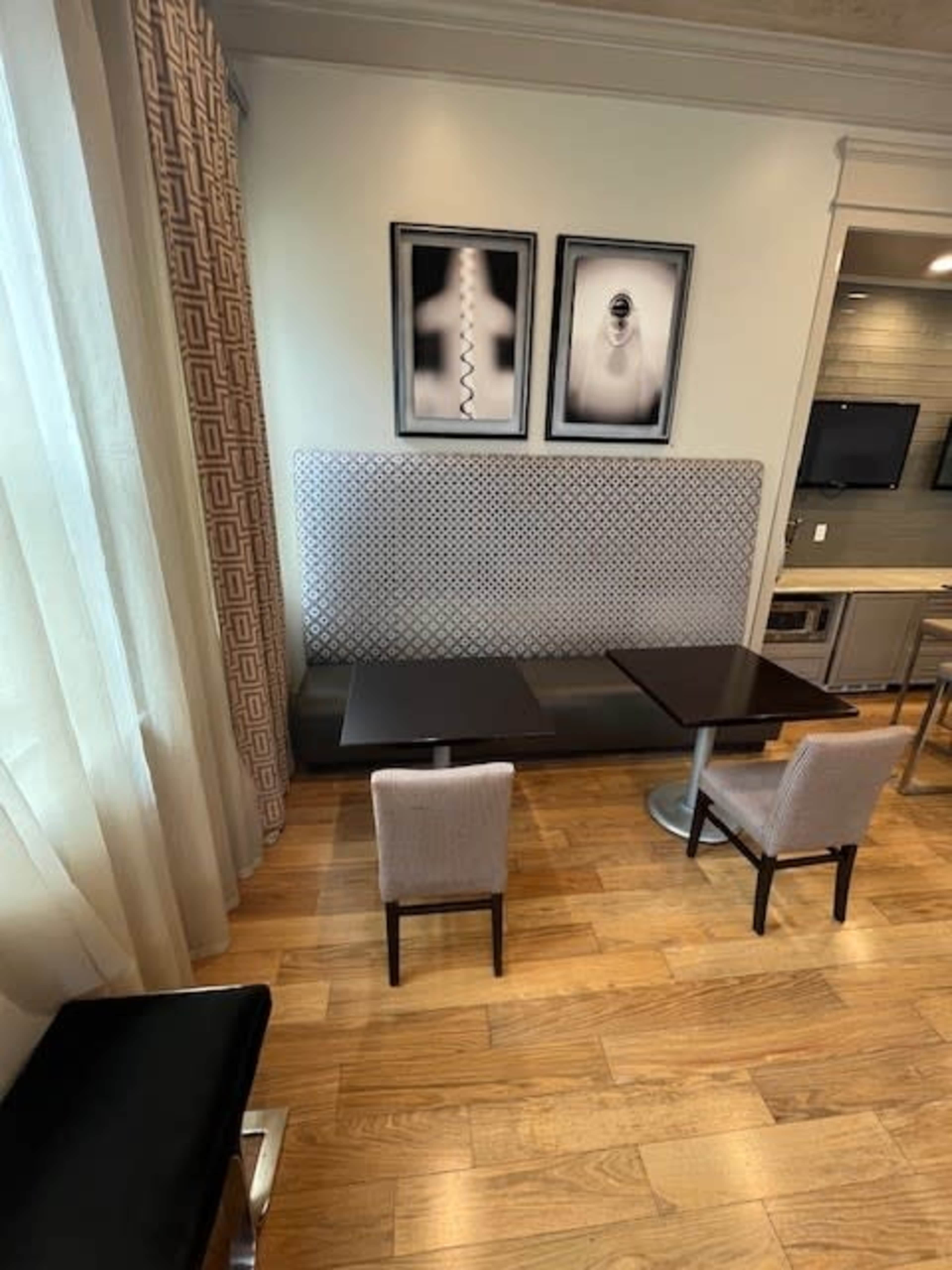 A dining area with a gray patterned booth, two small tables, and two chairs on a wooden floor, accompanied by two framed black-and-white photographs on the wall.