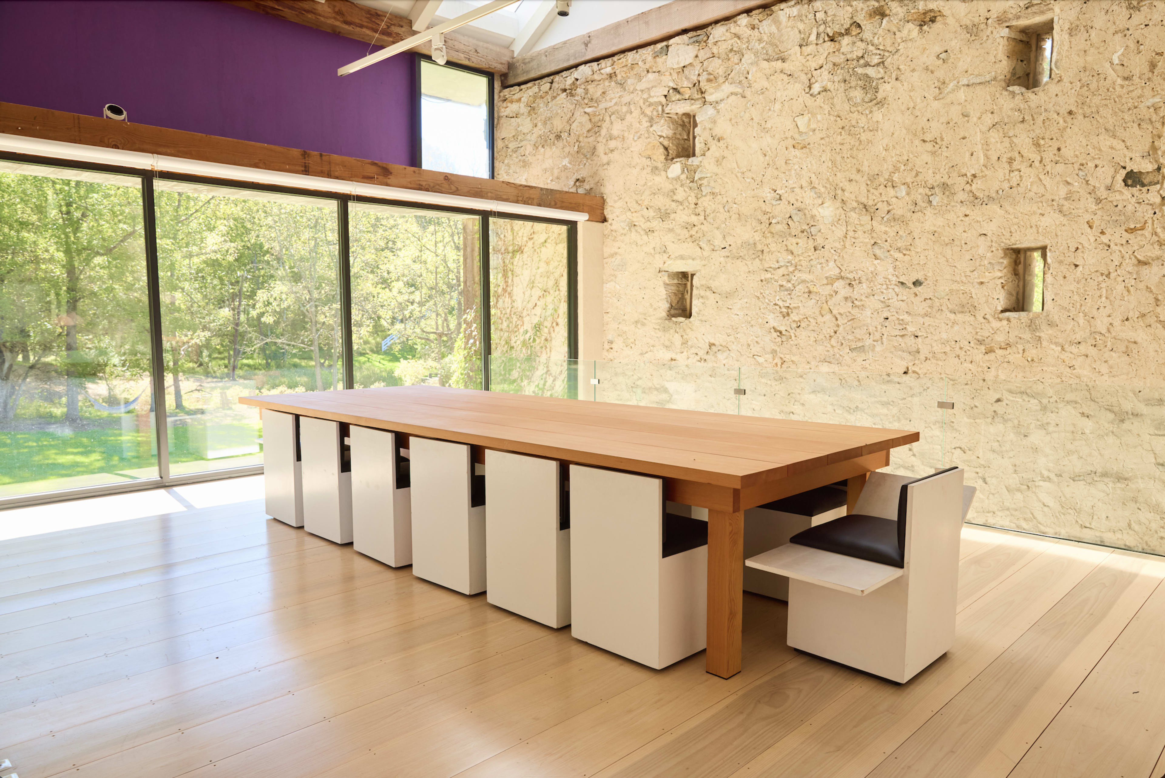 A large wooden table with white cube-shaped chairs is positioned in a modern room featuring a stone wall and large glass windows overlooking a green landscape.