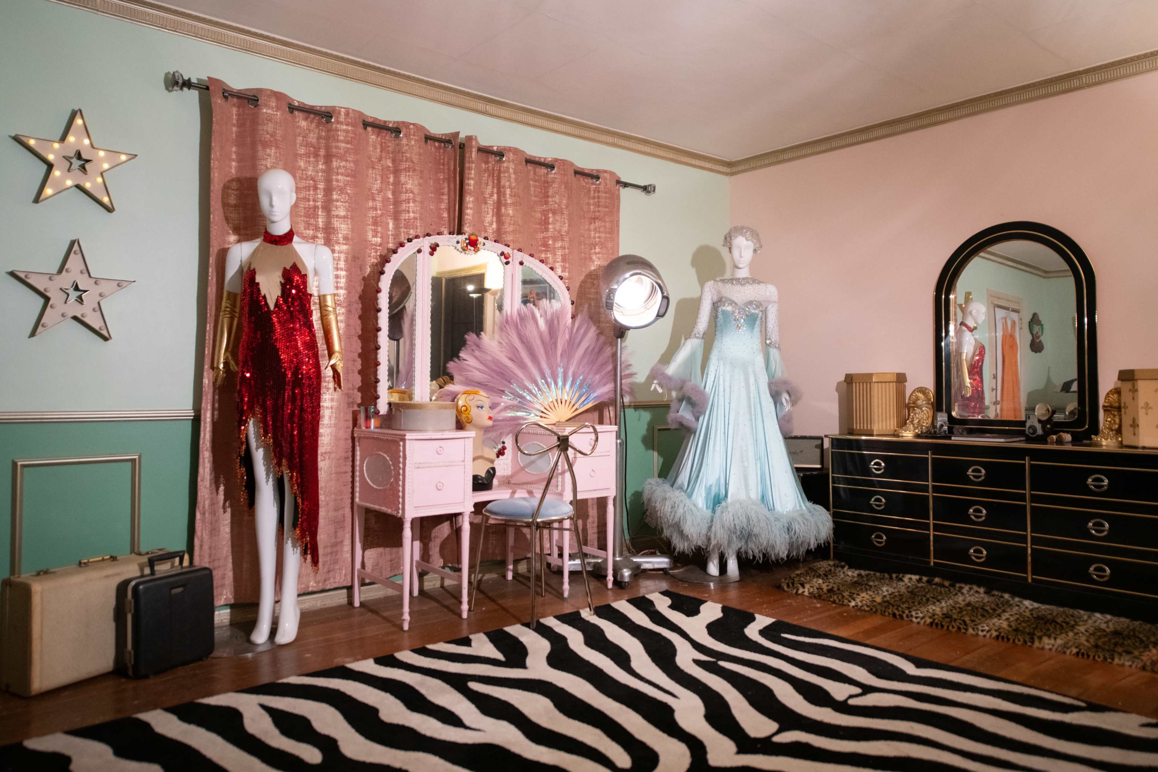 The image depicts a vintage-style room featuring mannequins dressed in elaborate costumes, a pink vanity with a mirror, and decorative elements like stars on the walls and a zebra-striped rug.