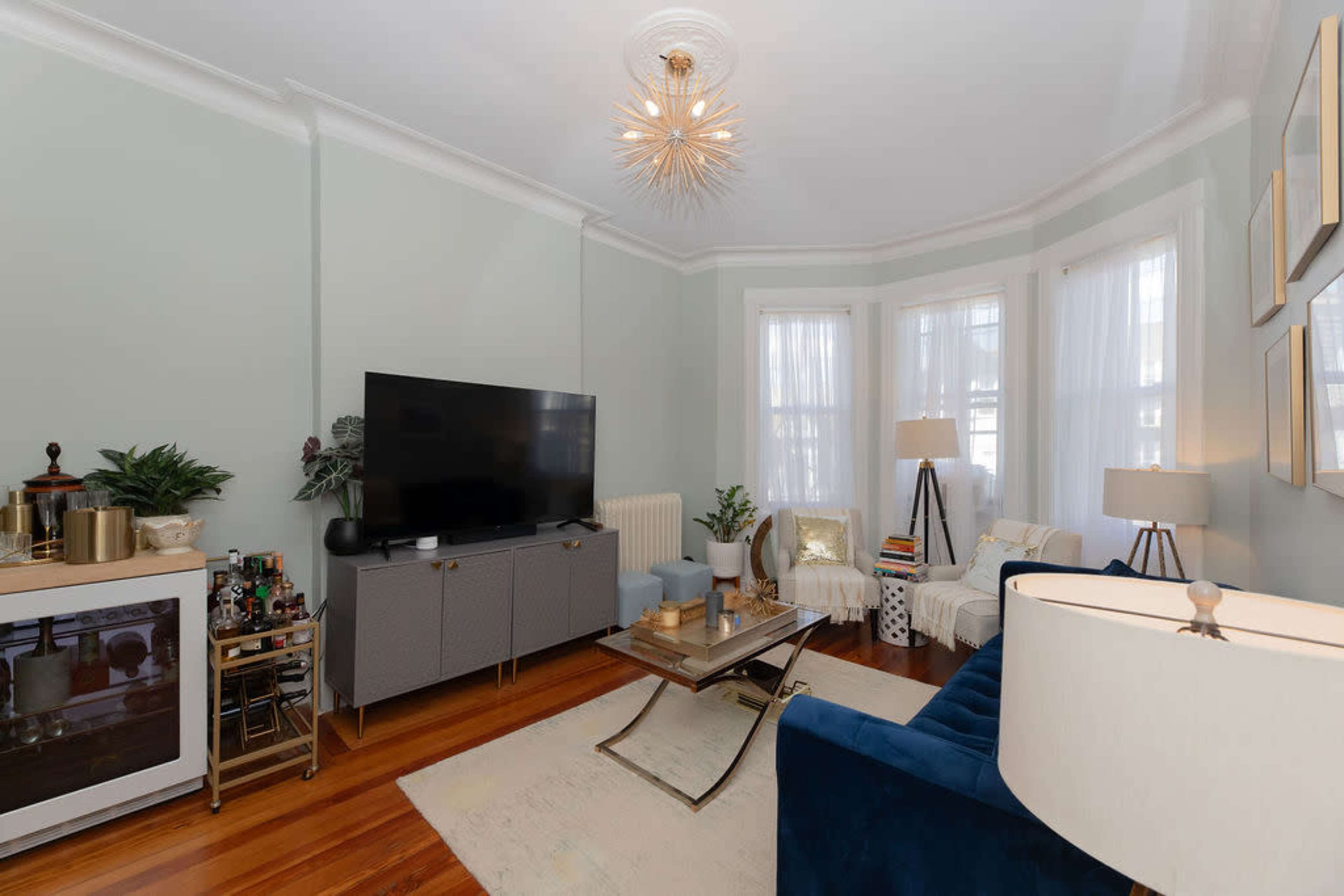 The living room features a large flat-screen television, a stylish blue couch, a decorative coffee table, and windows with sheer white curtains.