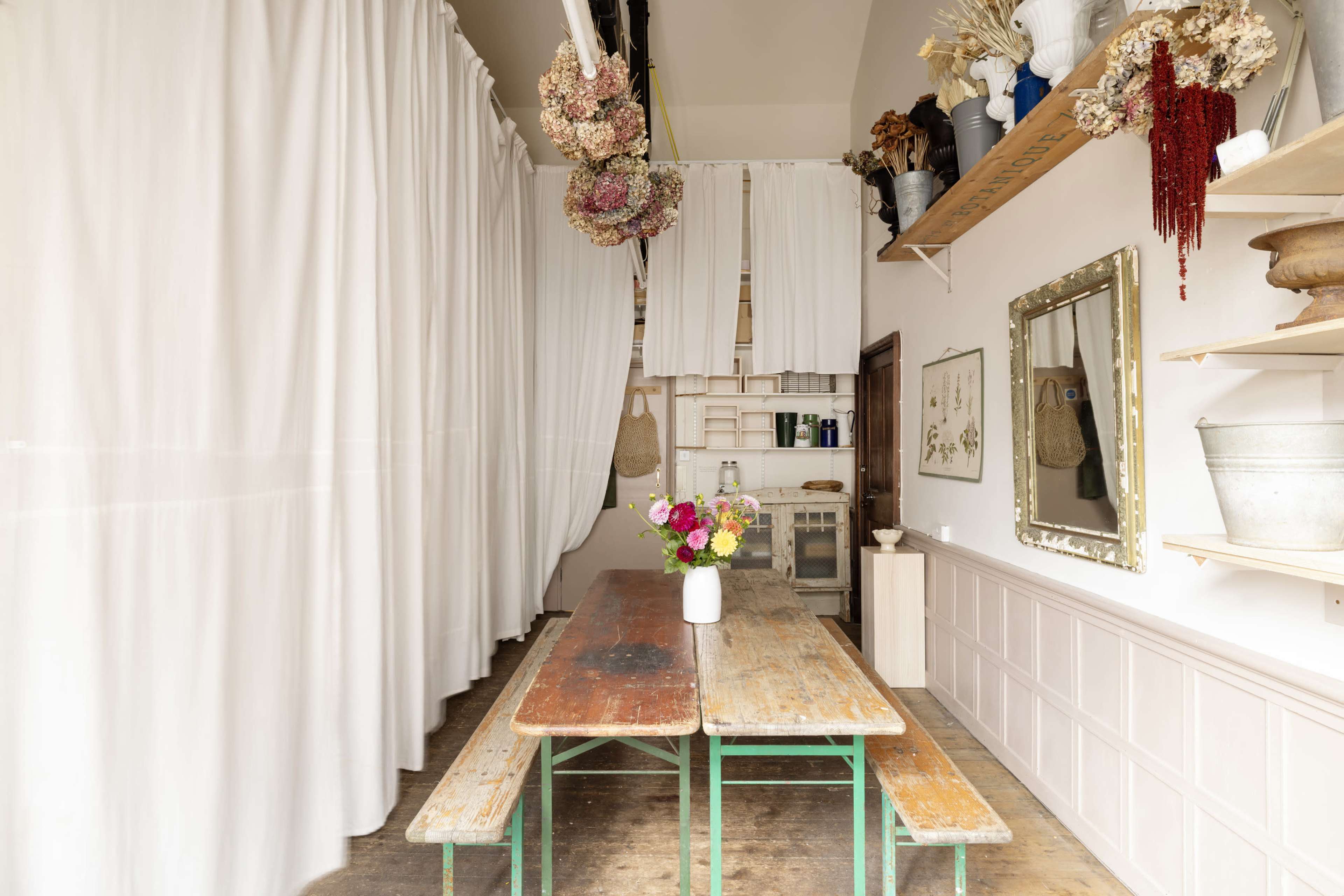 The image shows a rustic, light-filled interior featuring a long wooden table with a vase of flowers on it, surrounded by white curtains and shelves displaying decorative items.