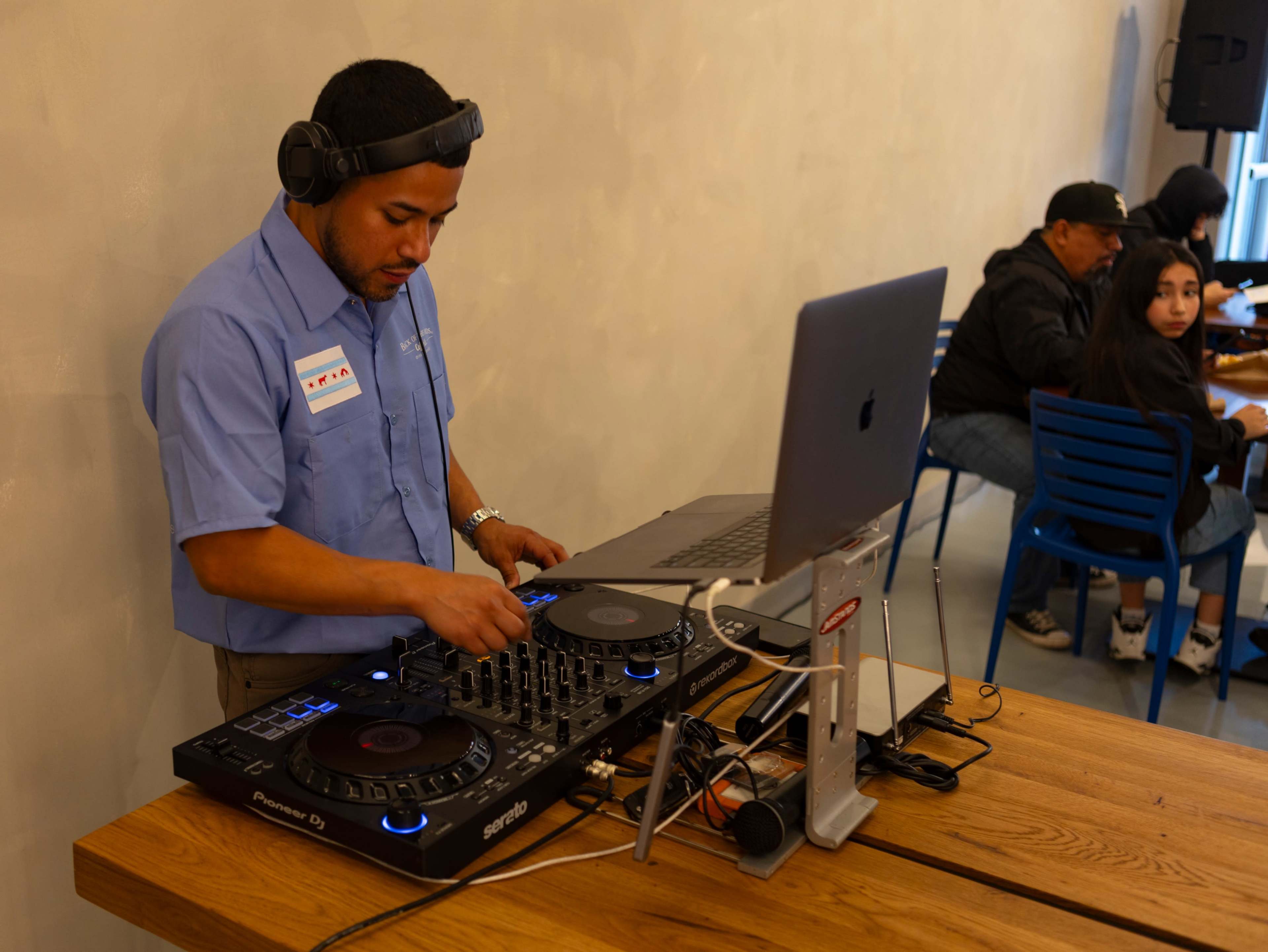 A DJ operates a mixing console and laptop while people sit and socialize in a cafe setting.