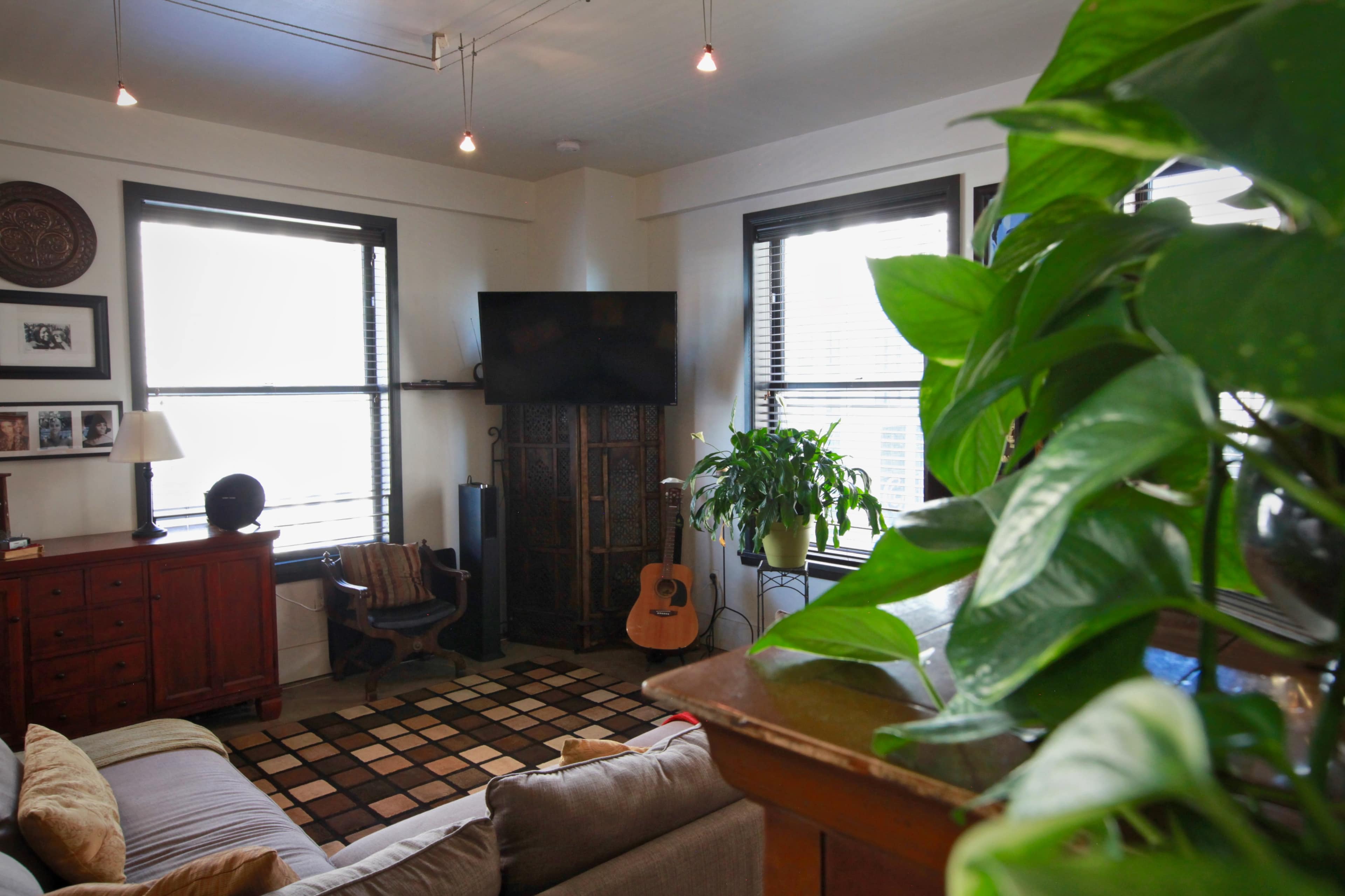 A cozy living room features a sofa, a wooden coffee table, an acoustic guitar, and large windows with potted plants.