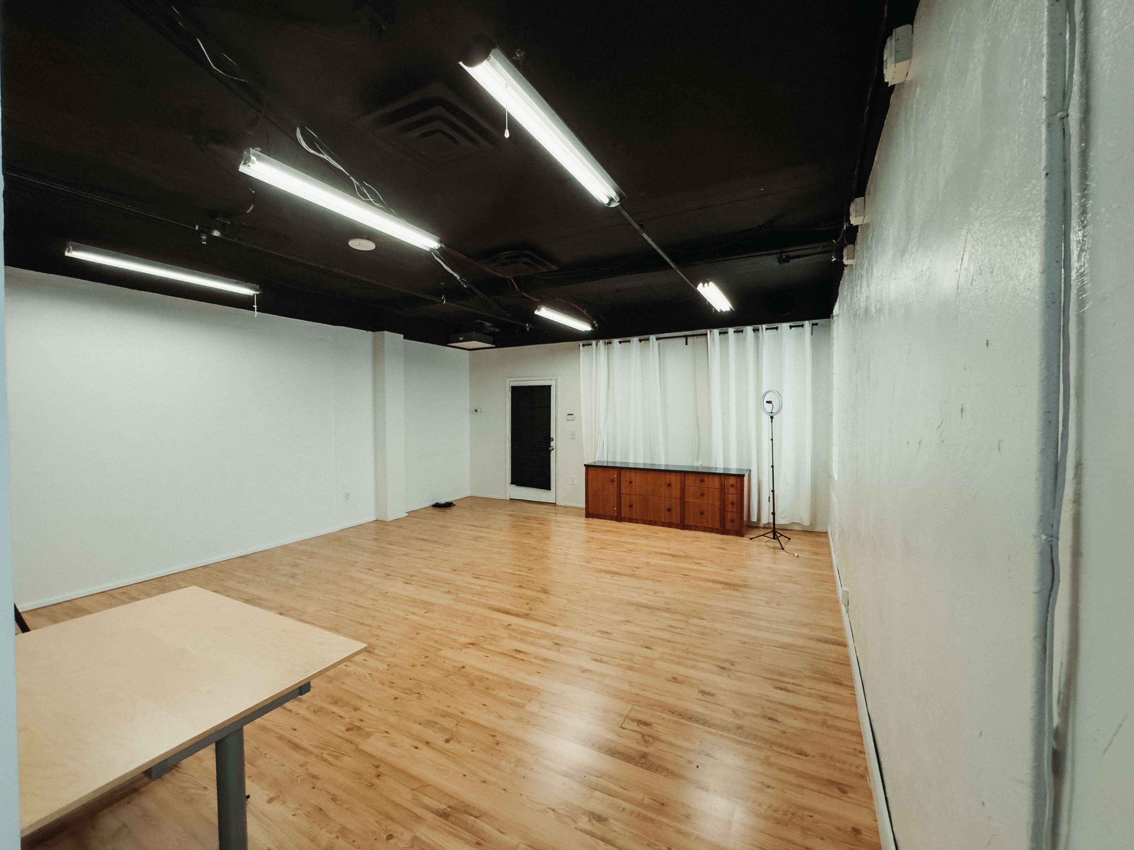 The image shows a sparse, well-lit studio space with wooden flooring, a white wall, and a wooden cabinet against the wall.