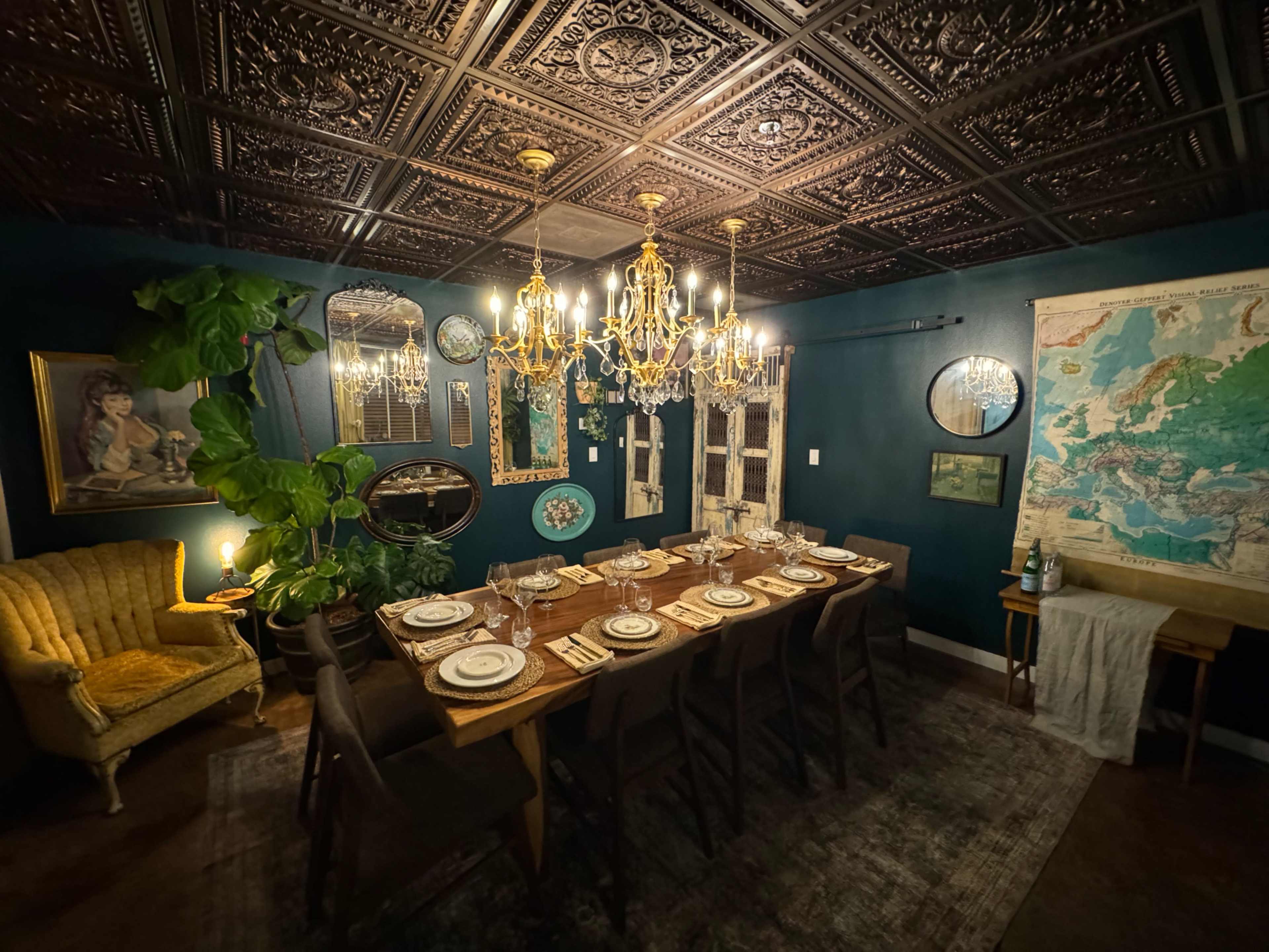 A large dining room features a long wooden table set for dinner, surrounded by dark chairs, with decorative wall items and a large chandelier illuminating the space.