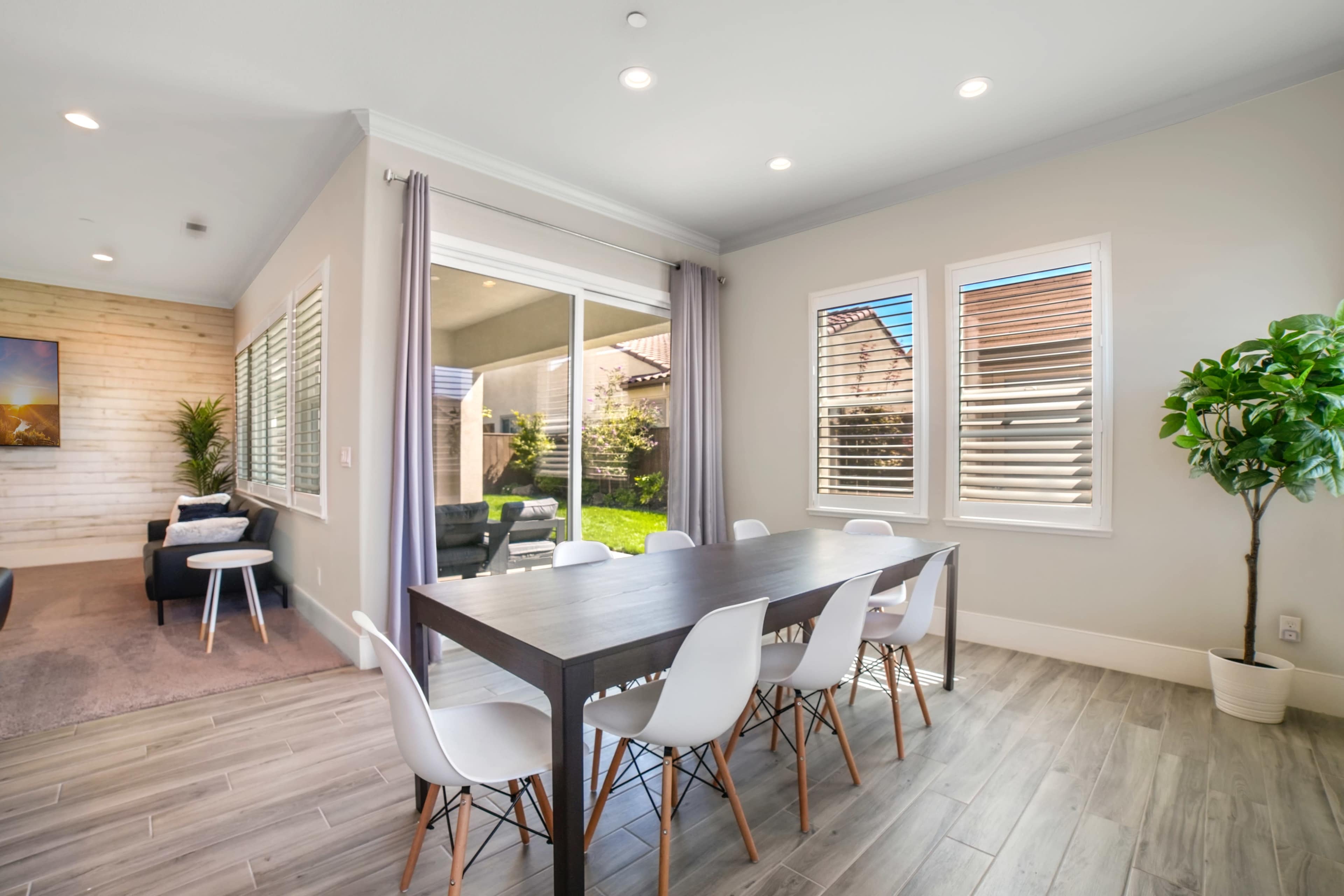 A modern dining area with a wooden table surrounded by white chairs, illuminated by natural light from nearby windows.