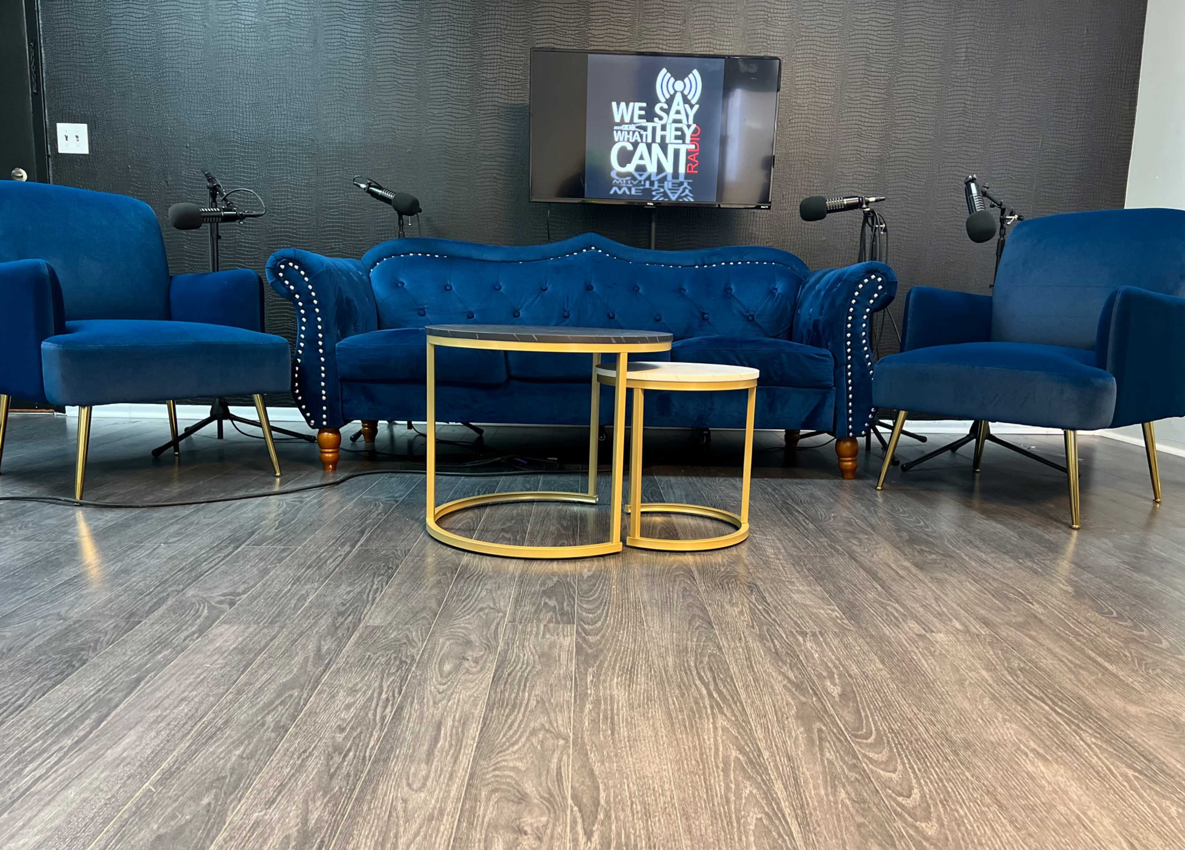 The image shows a modern seating area with a blue tufted couch and two matching armchairs, accompanied by two round gold coffee tables and a wall-mounted screen in the background.