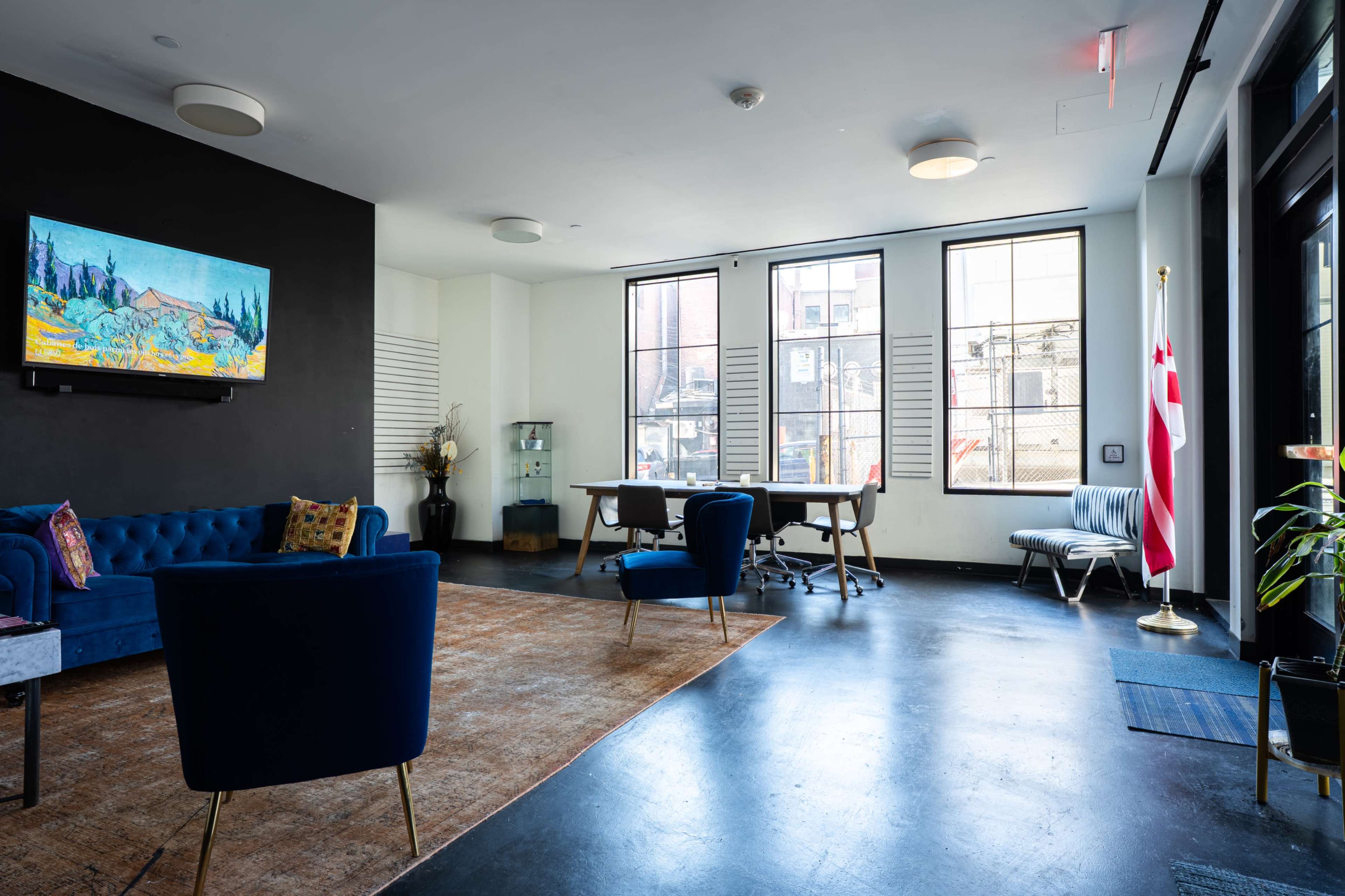 The image shows a modern office interior with a blue sofa, two blue chairs, a wooden desk, and large windows, featuring an artwork on the wall and a flag in the corner.