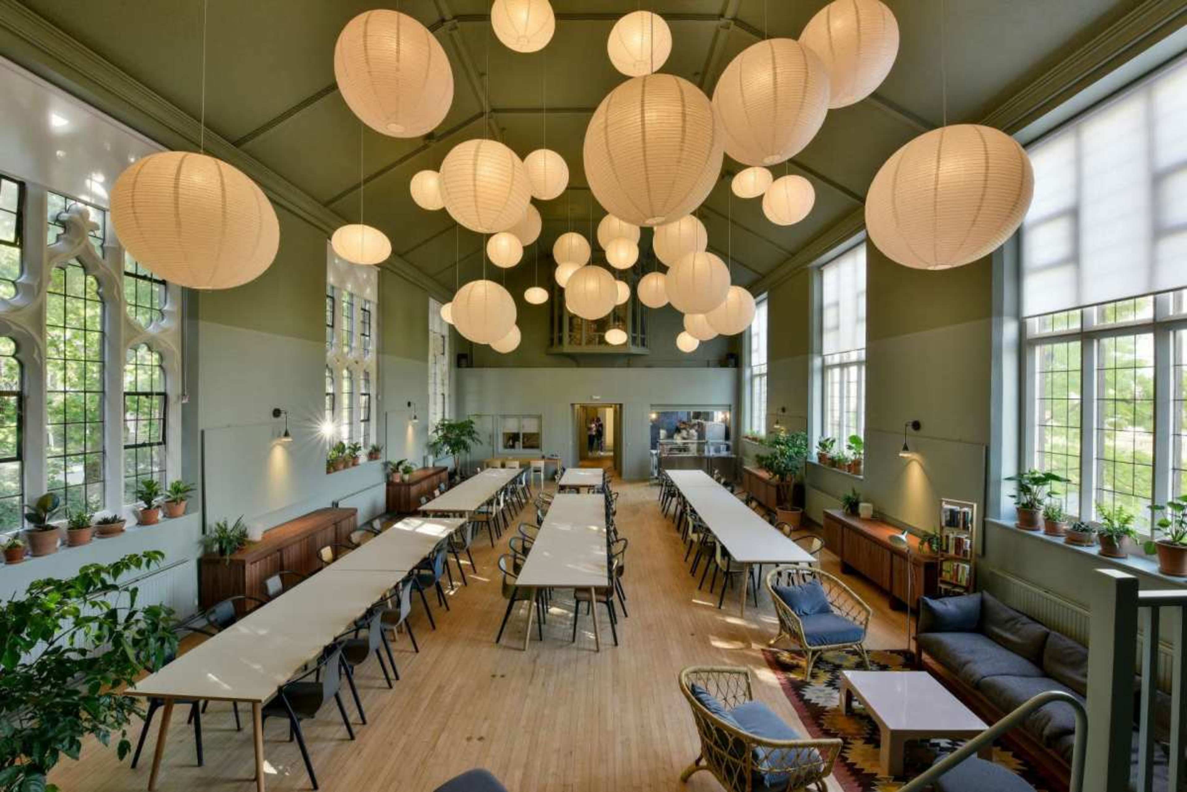 The image shows a spacious room with long tables arranged in rows, surrounded by large windows and decorated with hanging paper lanterns and potted plants.