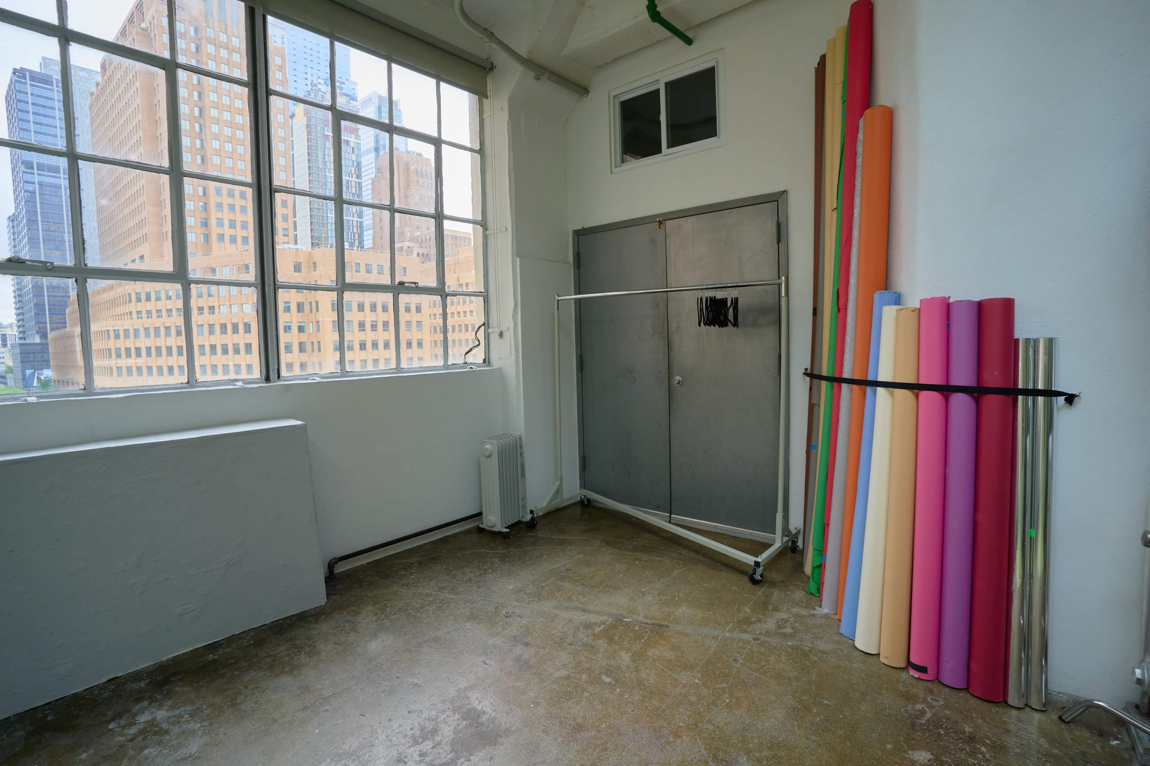 An empty studio space with large windows, a metal cabinet, and a rack holding rolls of colorful paper against a wall.