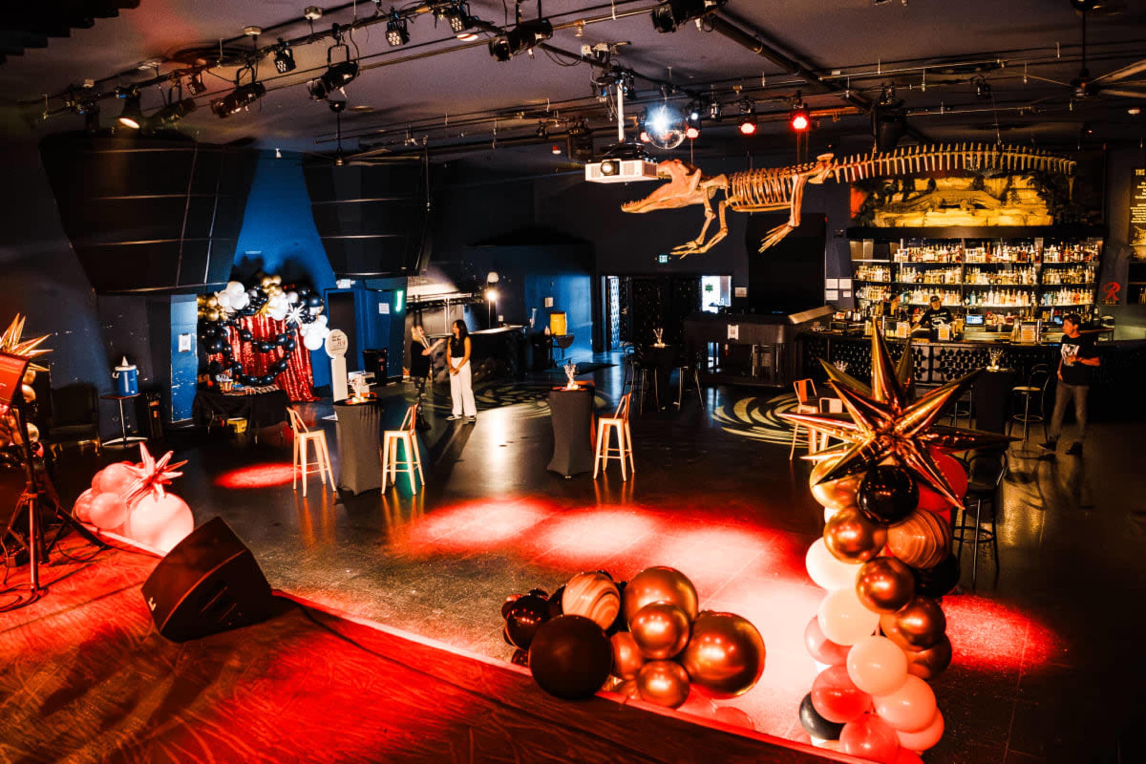 The image shows a large event space with a stage, bar area, and decorative balloon arrangements, highlighted by stage lighting and a dinosaur skeleton suspended from the ceiling.