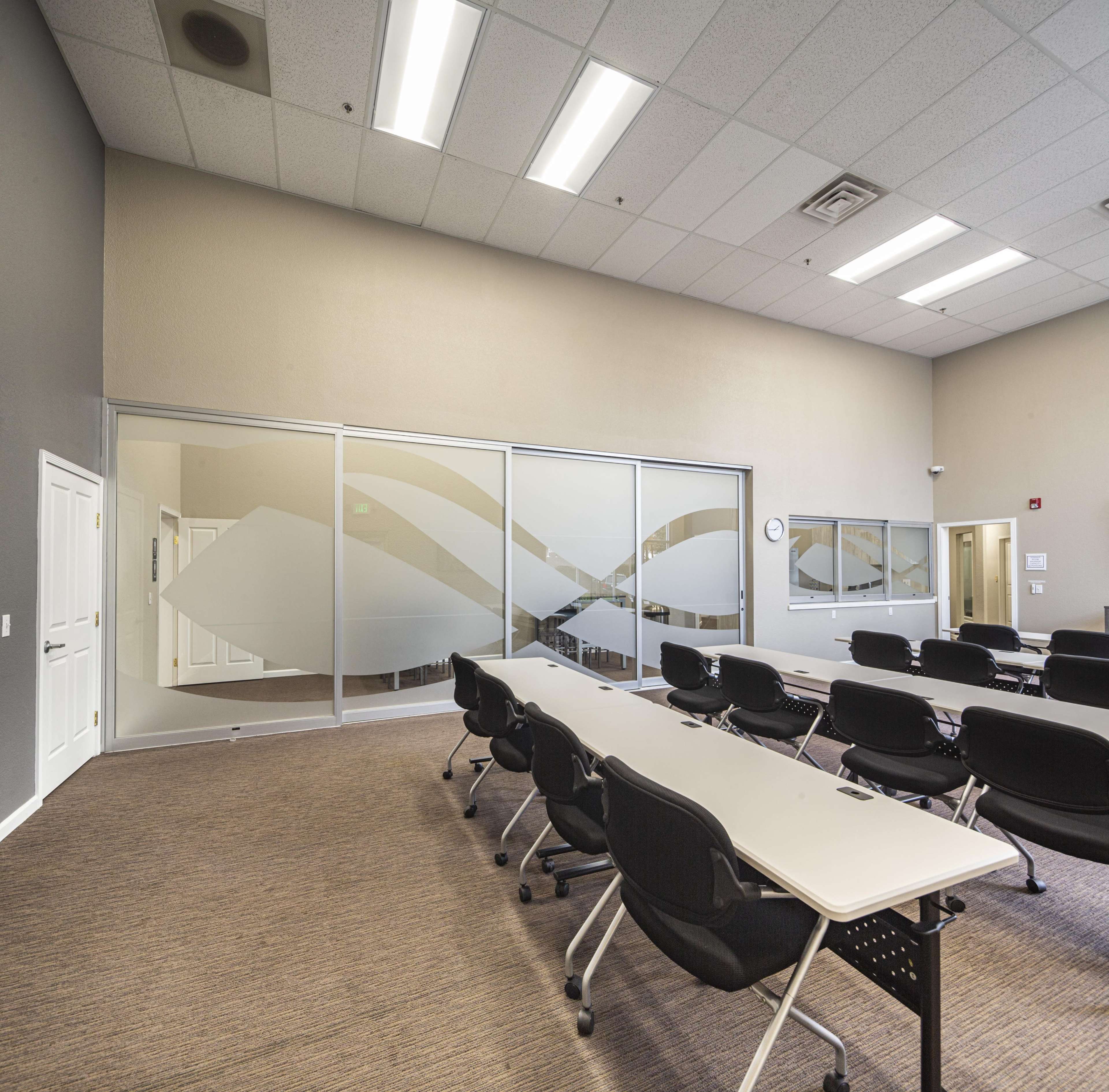 A spacious classroom features rows of black chairs and long white tables, with glass walls separating it from an adjoining area.