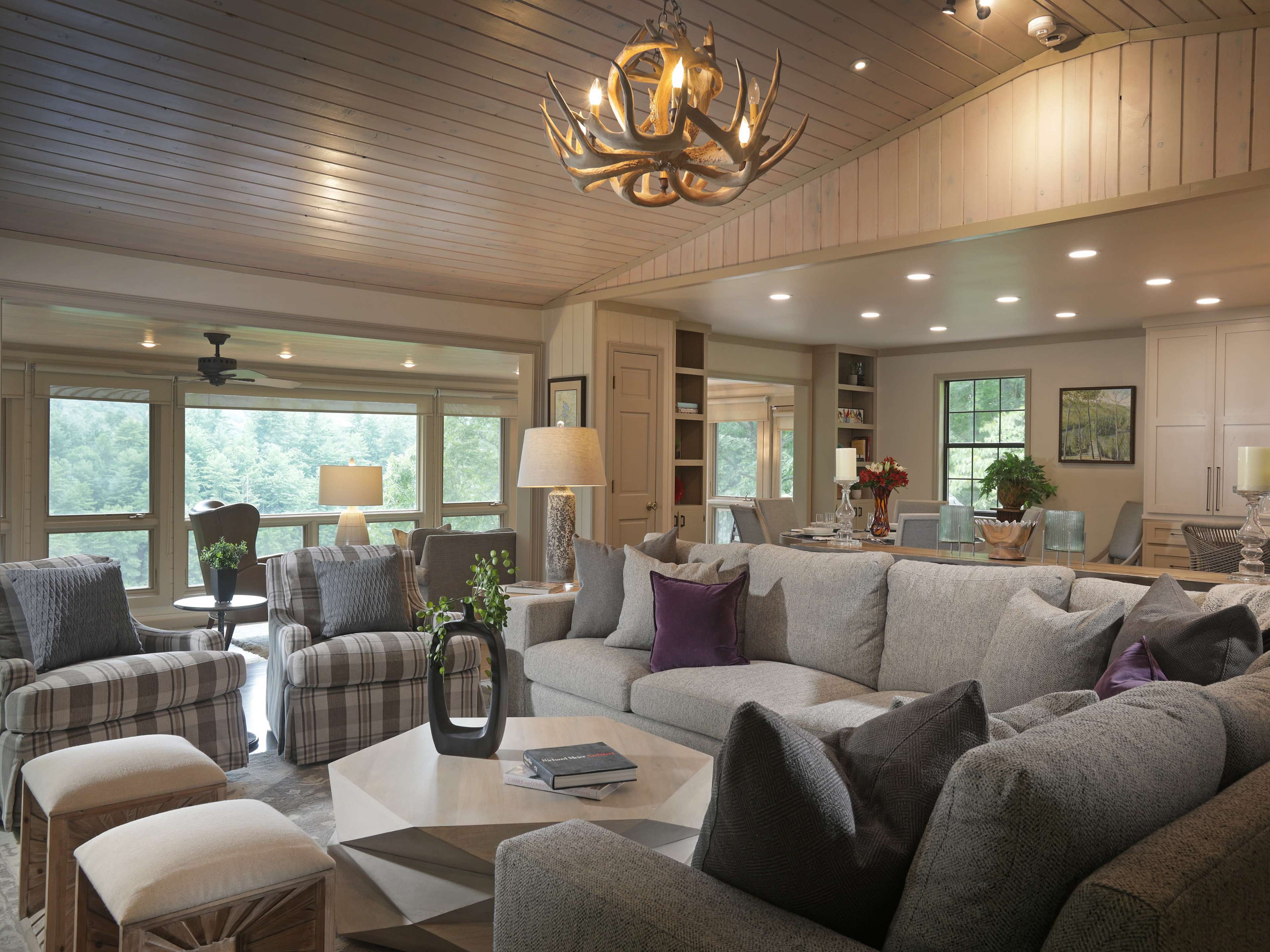 The living room features a spacious layout with a large sectional sofa, a coffee table, various seating options, and large windows overlooking greenery.