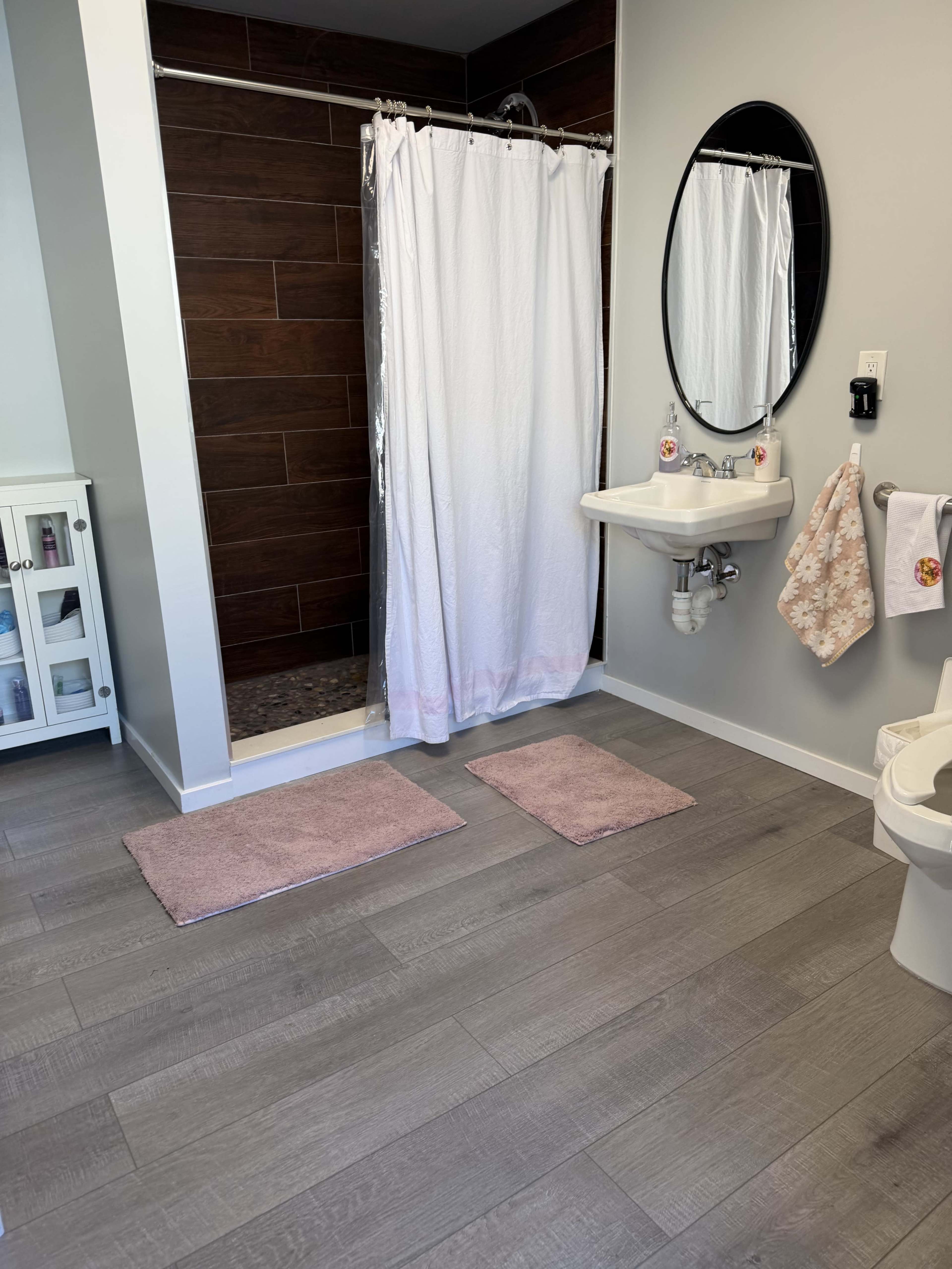 The image shows a bathroom featuring a shower with a white curtain, a sink with a circular mirror above it, and light-colored flooring with two pink bath mats.