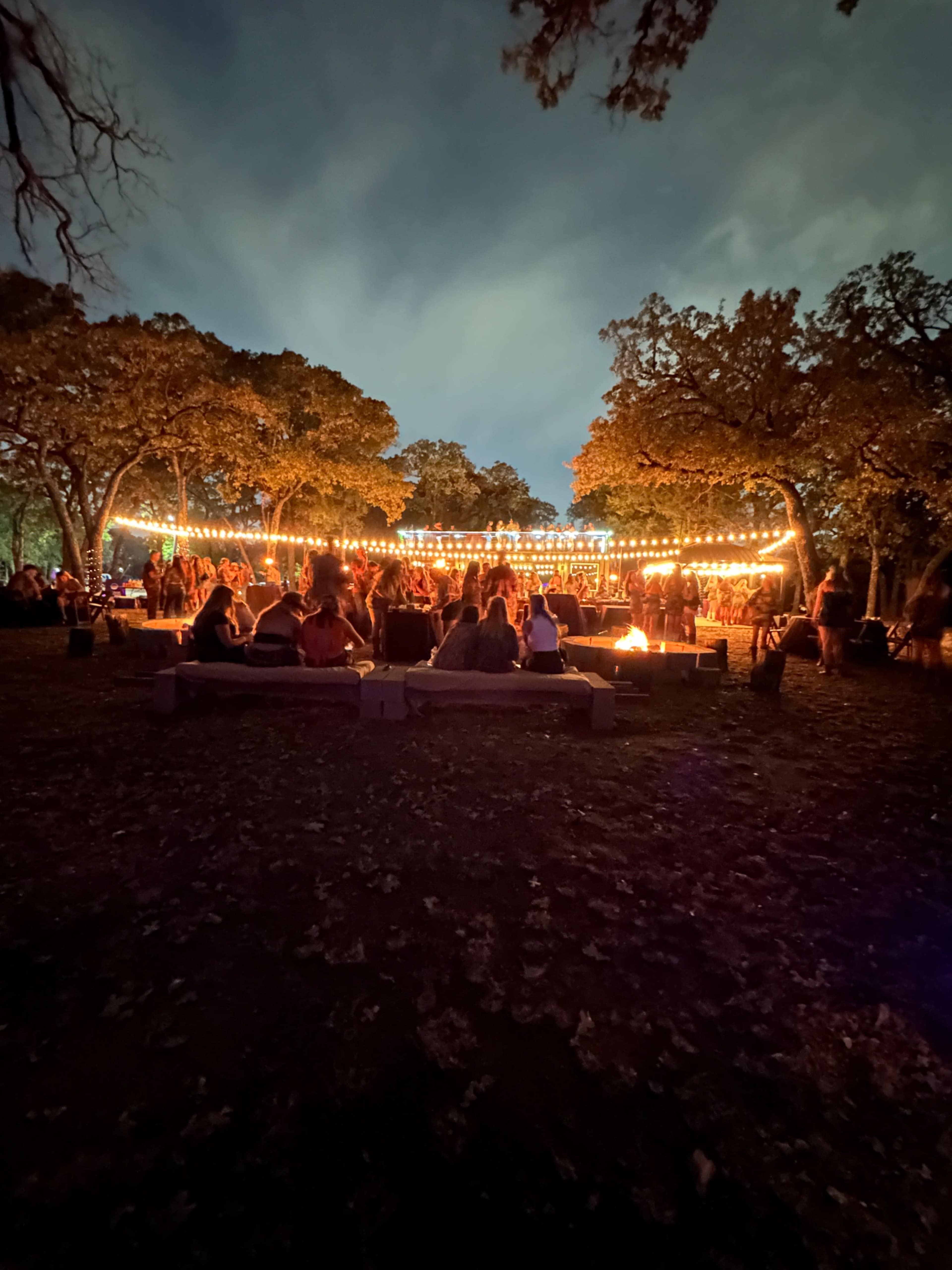 A group of people gathers around a campfire, surrounded by trees and illuminated by string lights in the evening.