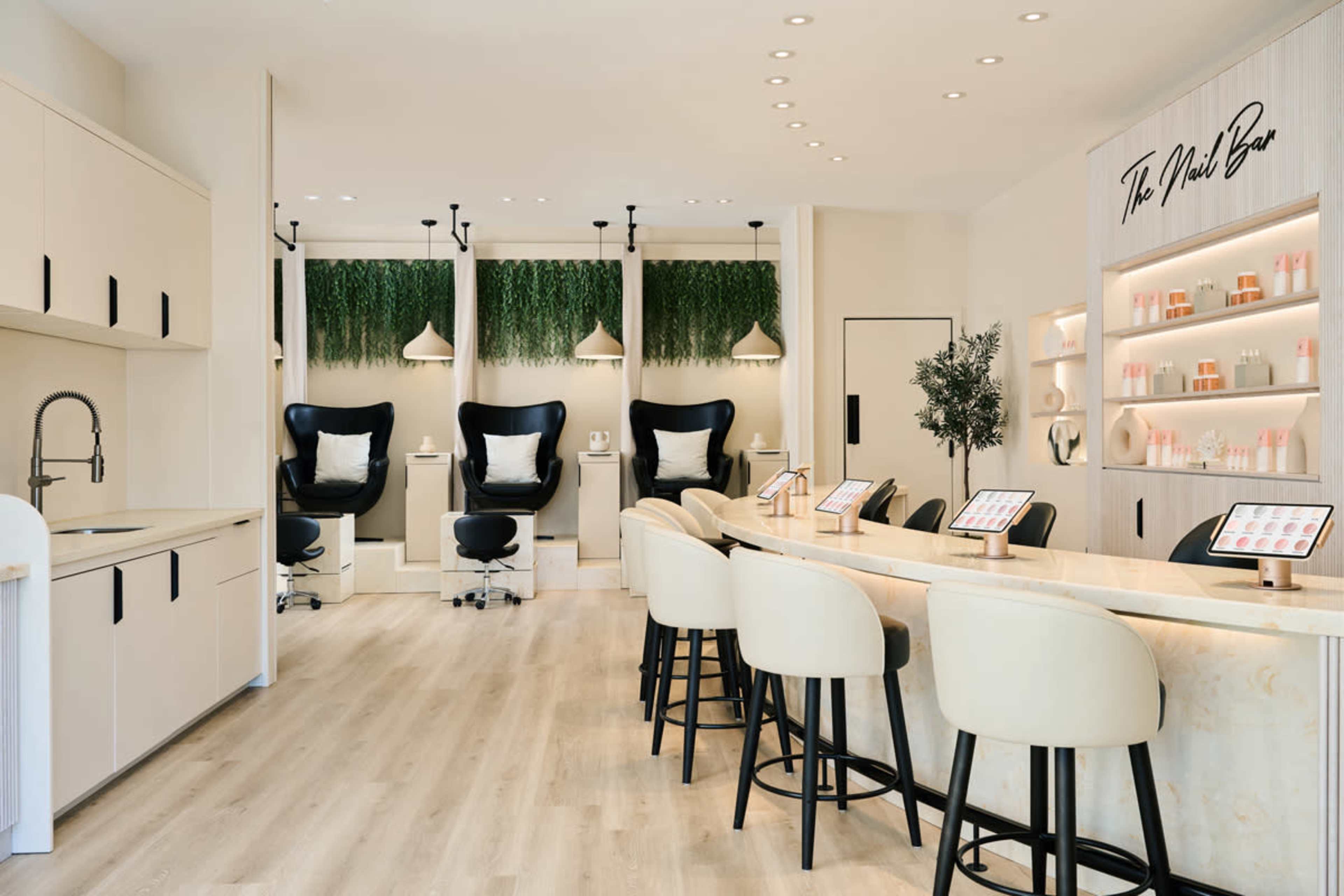 The image showcases a modern beauty salon interior featuring a long marble counter with bar seating, three treatment chairs, and greenery accents on the walls.