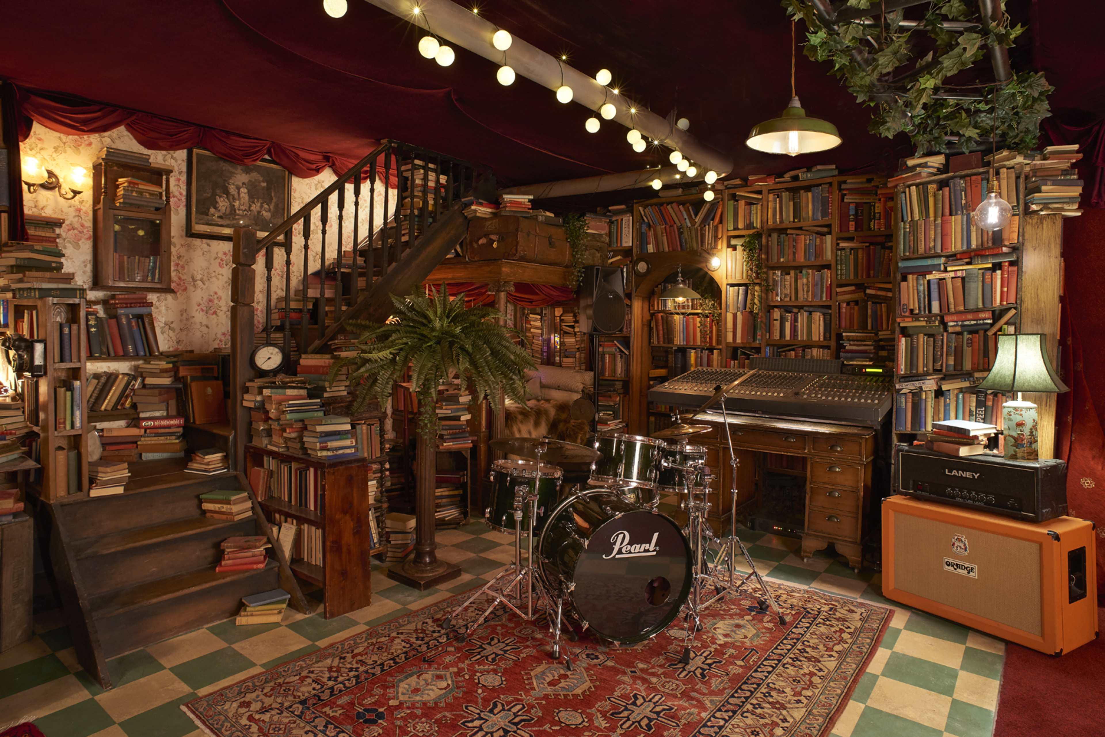 The image shows a cozy room filled with bookshelves stacked with books, featuring a drum set, a sound mixer, and a decorative rug, all set beneath warm lighting.