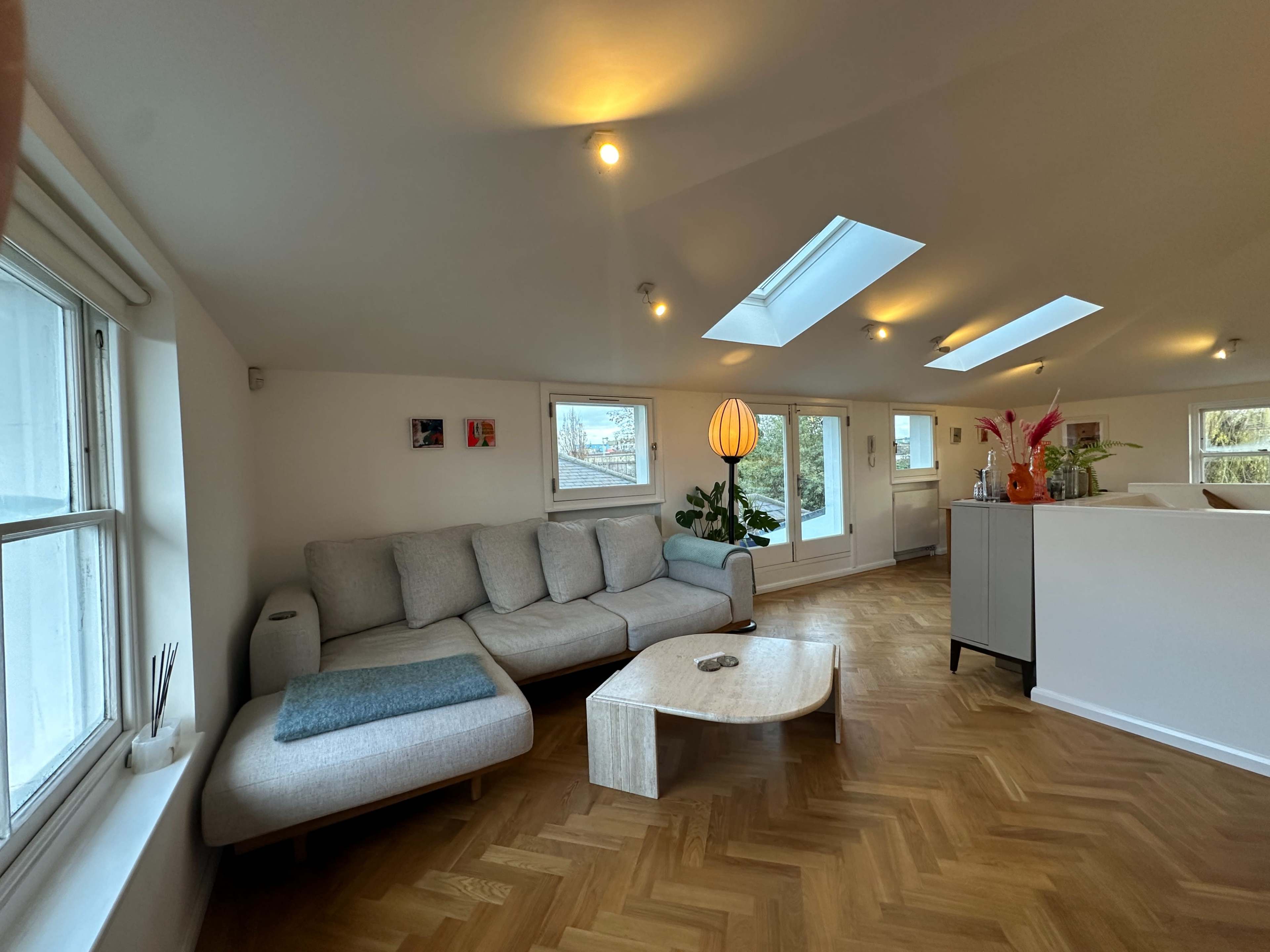 The image shows a modern, minimalistic living room with a light gray sectional sofa, a wooden coffee table, and large windows allowing natural light to enter.