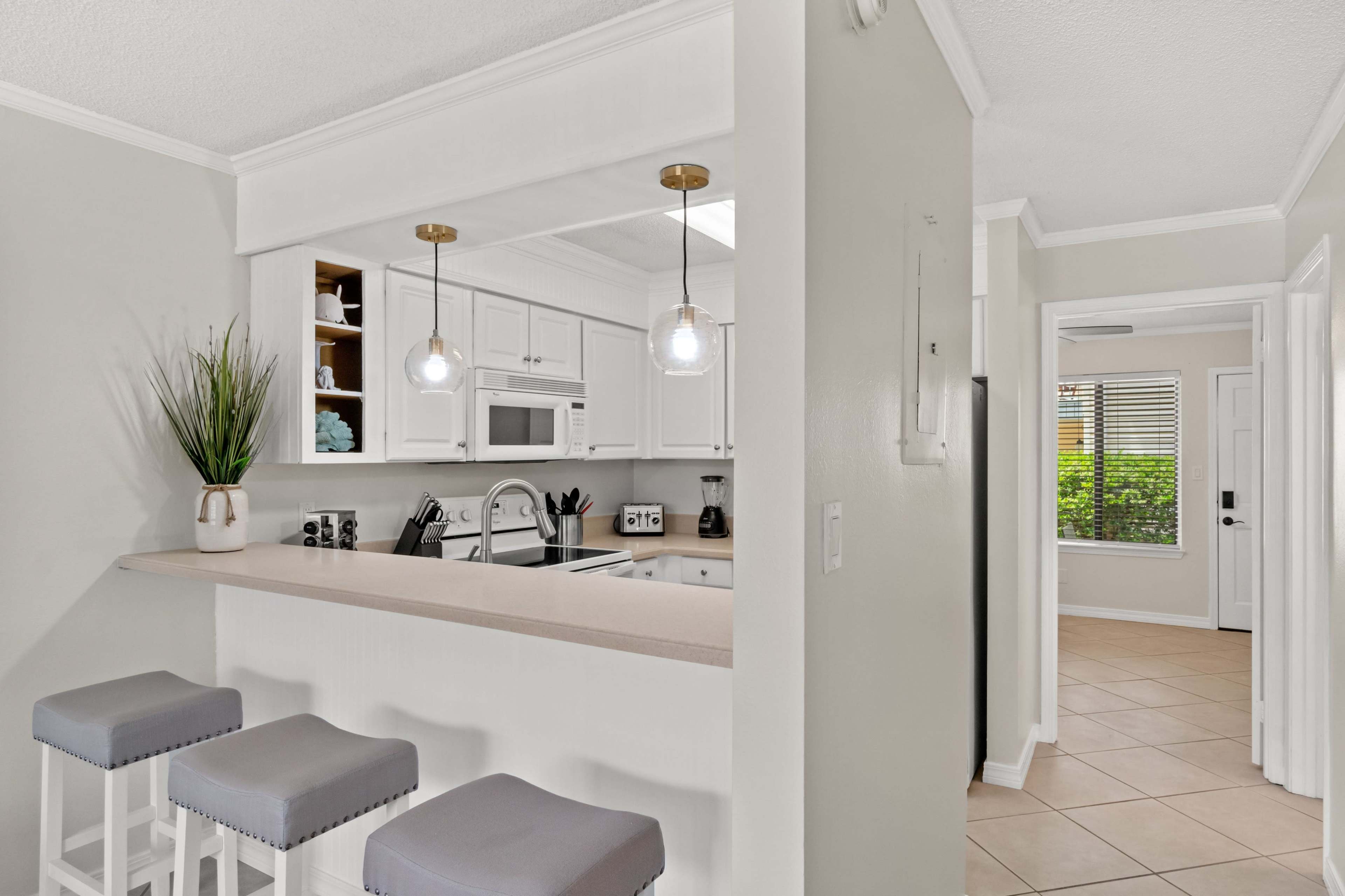A kitchen area features a breakfast bar with three stools, white cabinetry, and a view toward a hallway with a door leading outside.