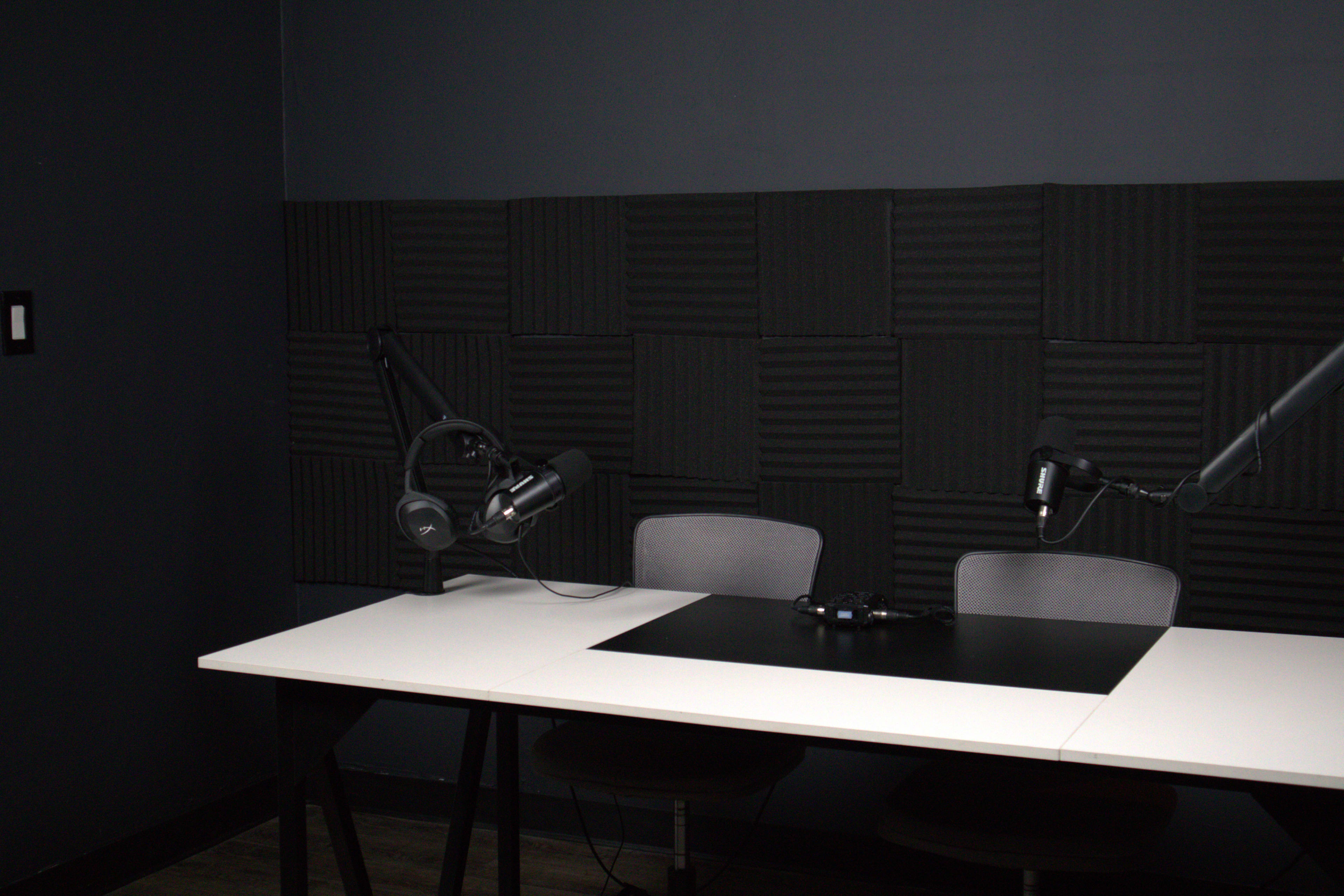 A podcast recording studio with two microphones mounted on adjustable arms positioned over a table and black soundproofing panels on the wall.