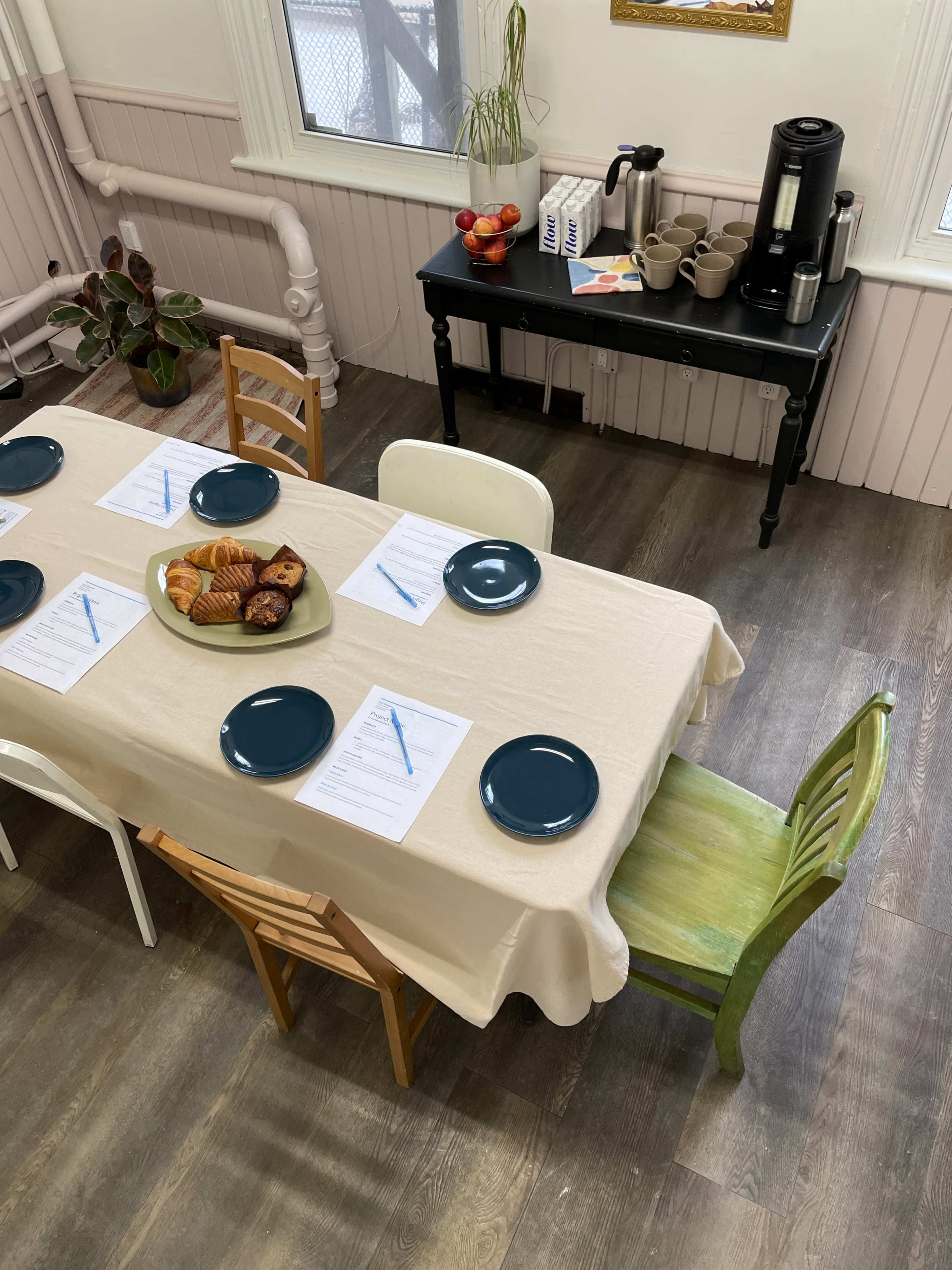 A large, light-colored table is set with blue plates and notepads, surrounded by mismatched chairs, with a platter of pastries and a coffee station in the background.
