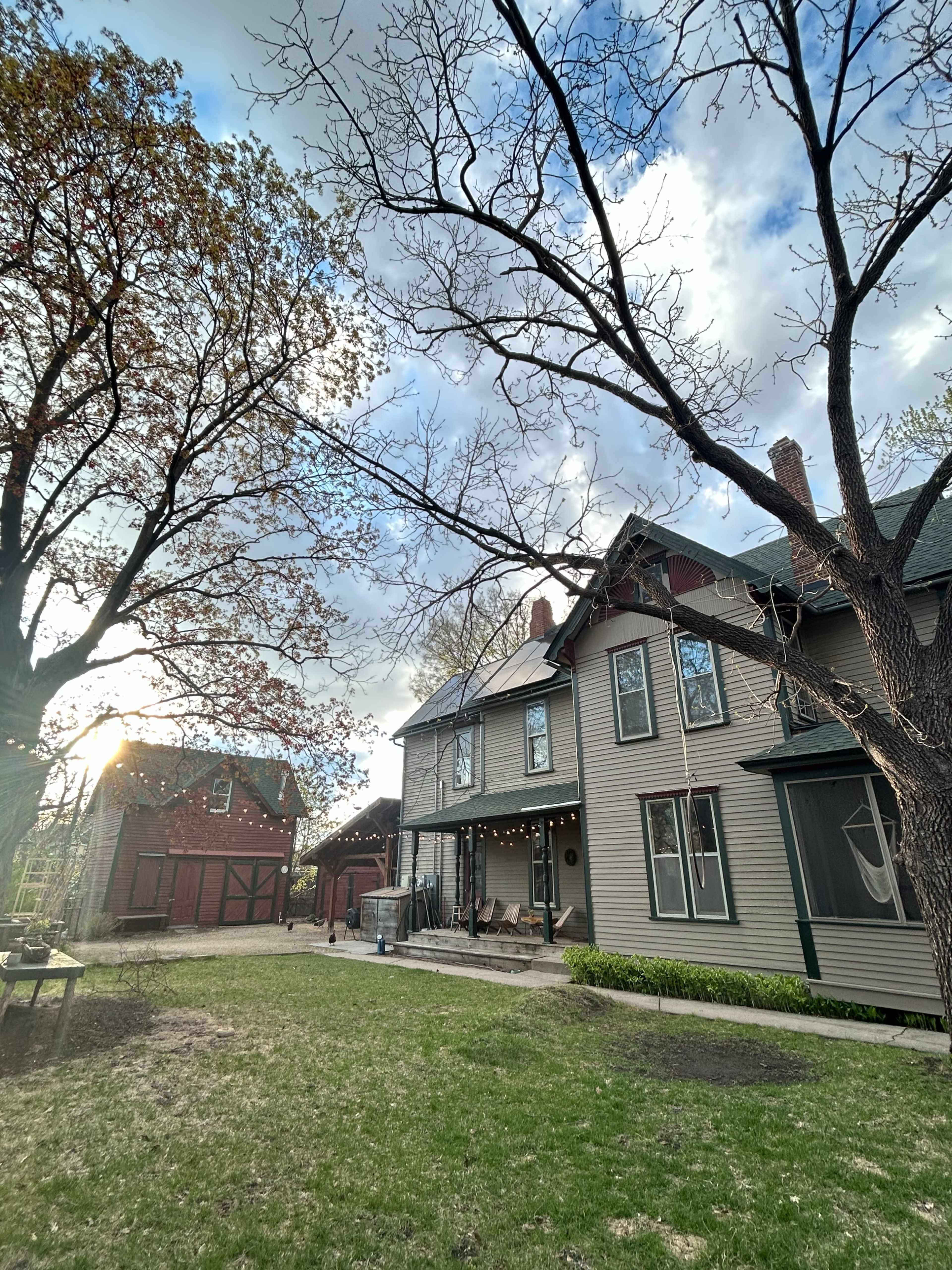A gray two-story house with a porch and string lights is situated beside a large tree in a grassy yard, with another building visible in the background.