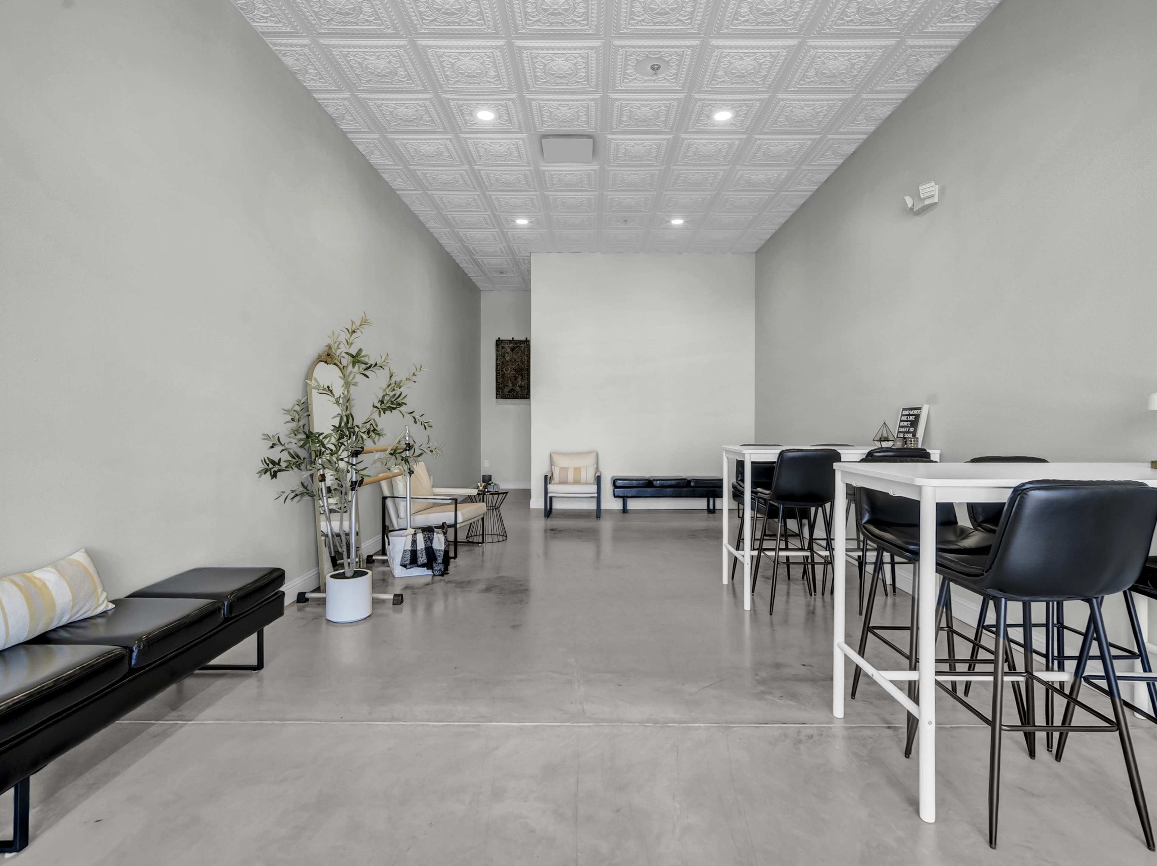 The image shows a minimalist interior space with concrete floors, white walls, a few tables and chairs, and a small plant.