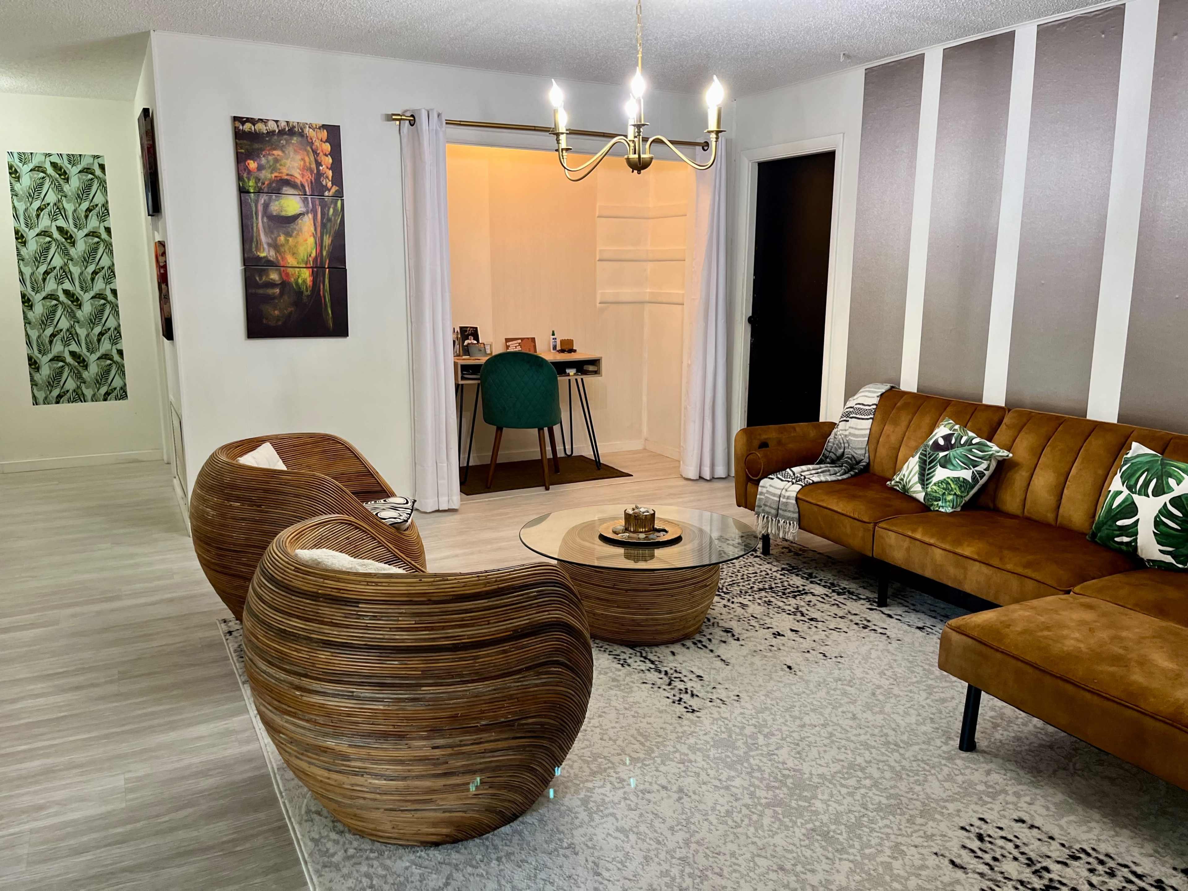A modern living room features a brown leather sofa, two woven chairs, a glass coffee table, and a workspace area in the background with a green chair.