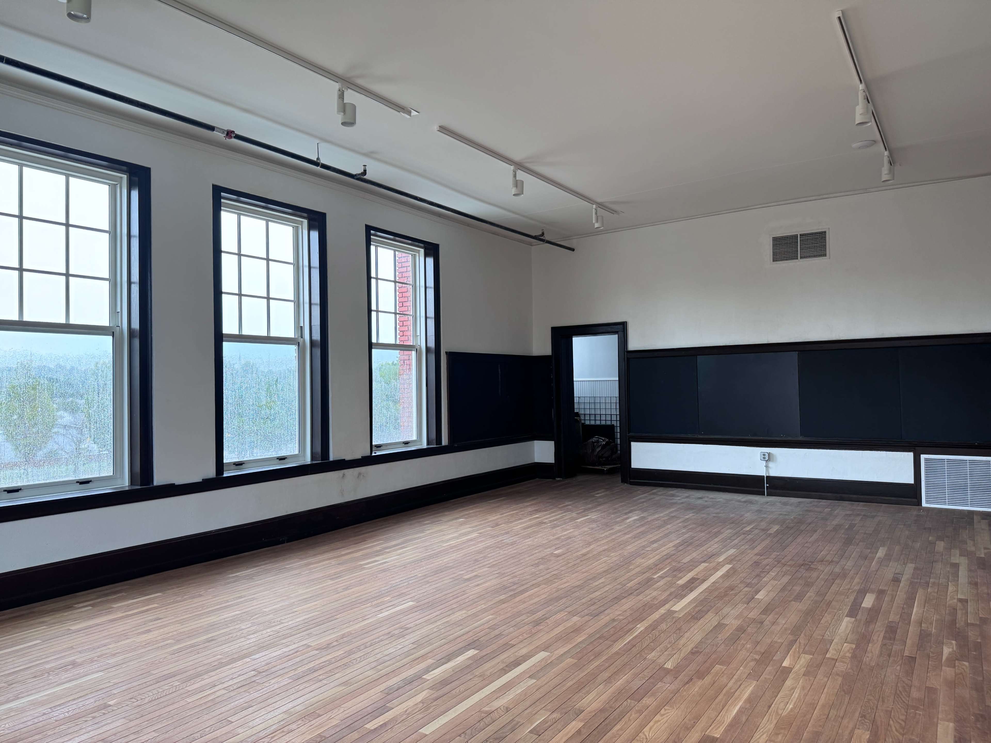 The image shows an empty room with large windows, wooden flooring, and black and white walls.