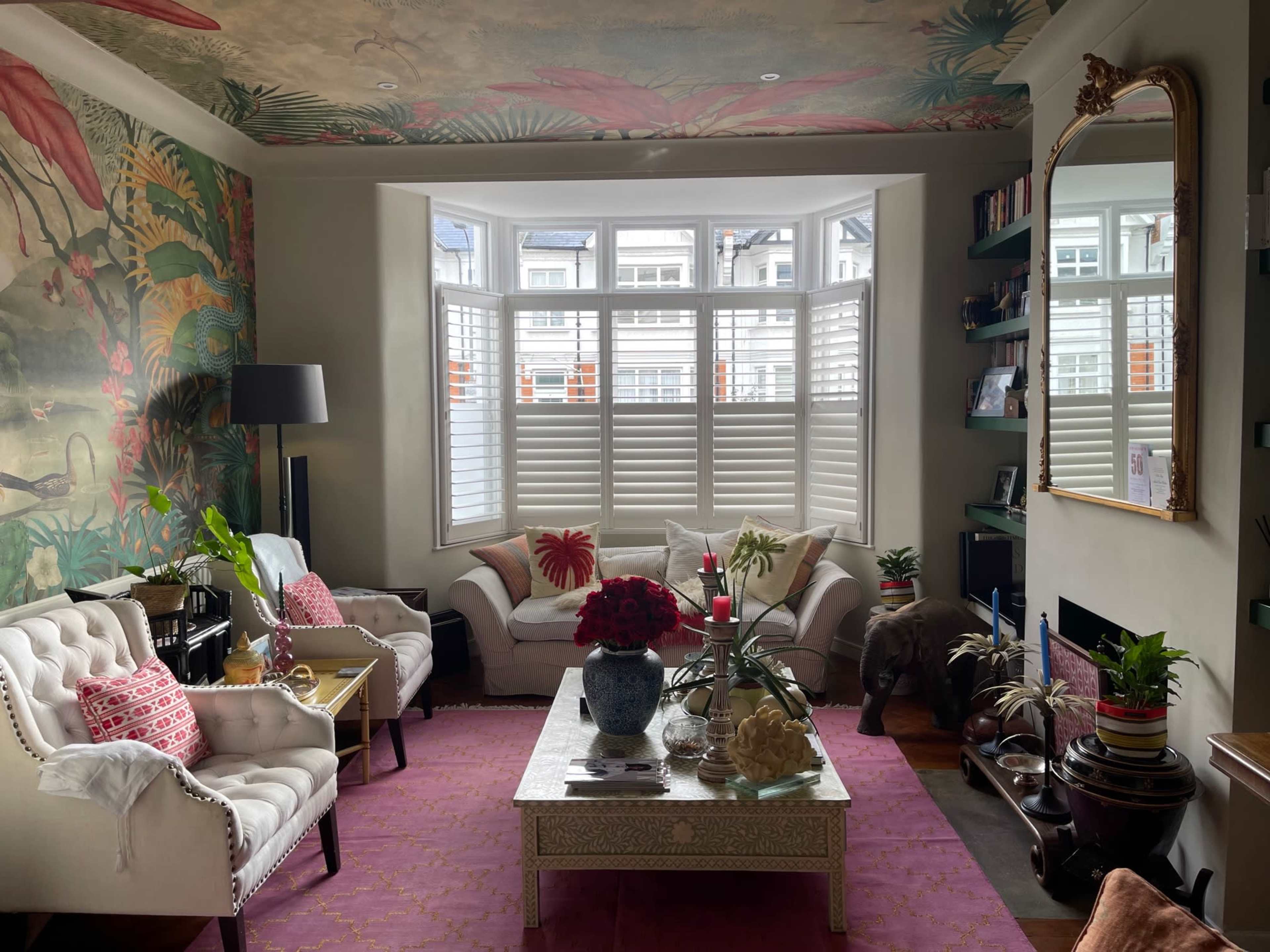 A living room features floral wallpaper, a bay window with plantation shutters, a white sofa, and a coffee table adorned with decorative items.