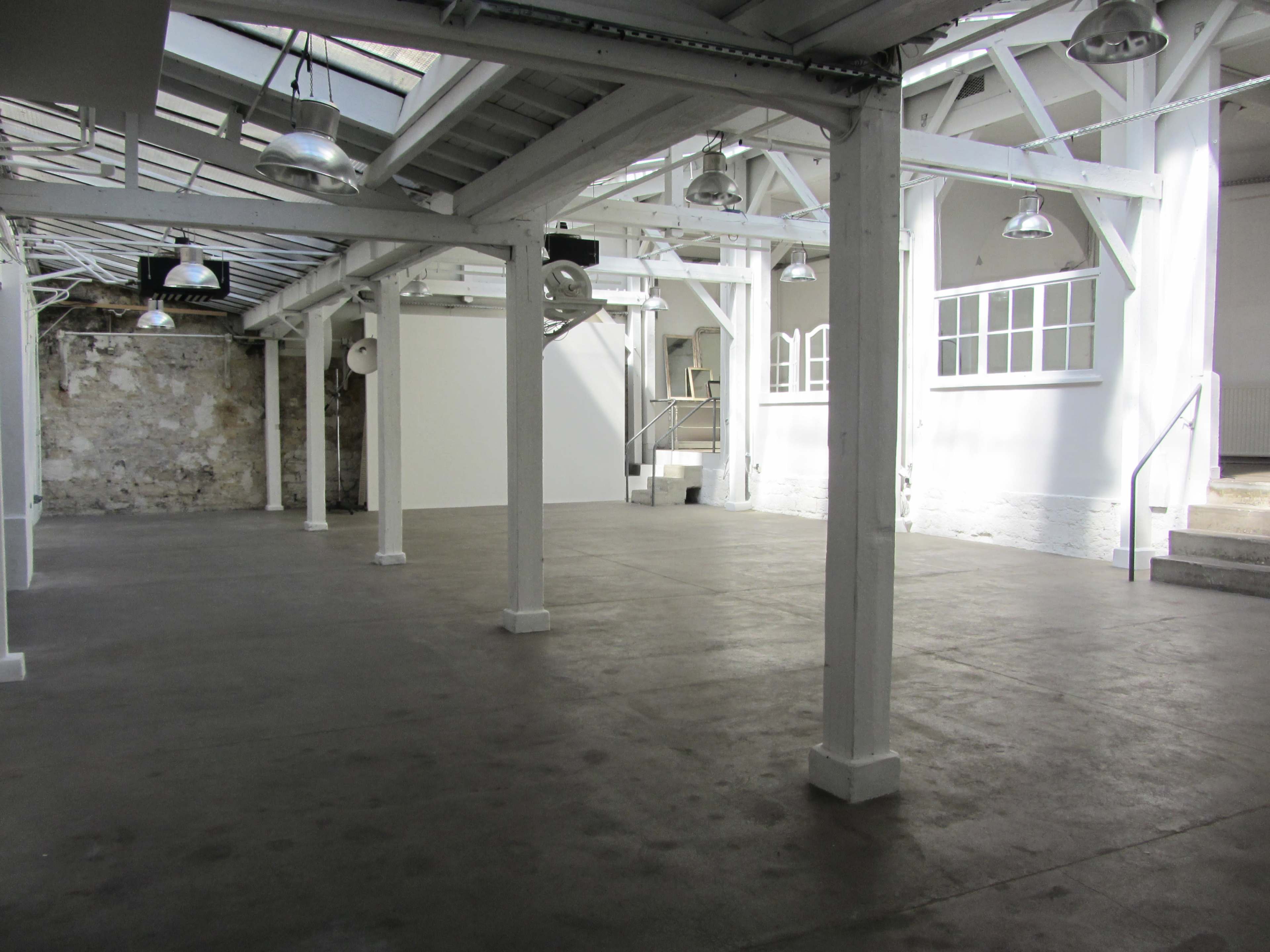 The image shows an empty, well-lit industrial space with white walls, concrete flooring, and exposed wooden beams.