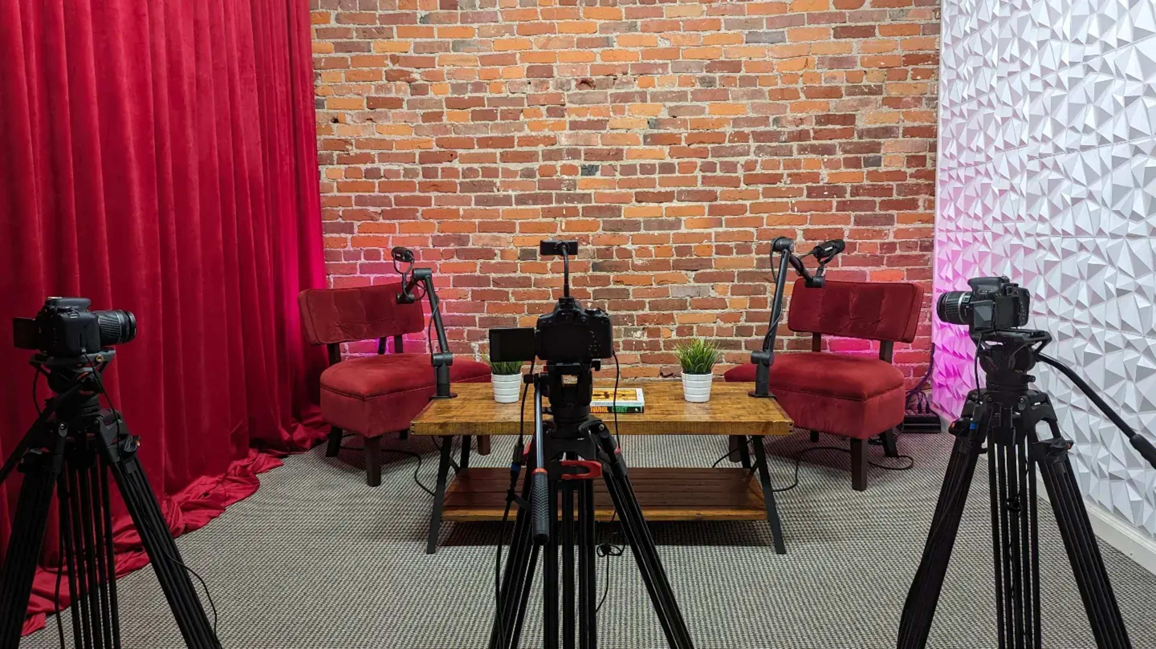 A video recording setup featuring two red chairs and a wooden table, surrounded by three tripods holding cameras, against a brick wall backdrop.