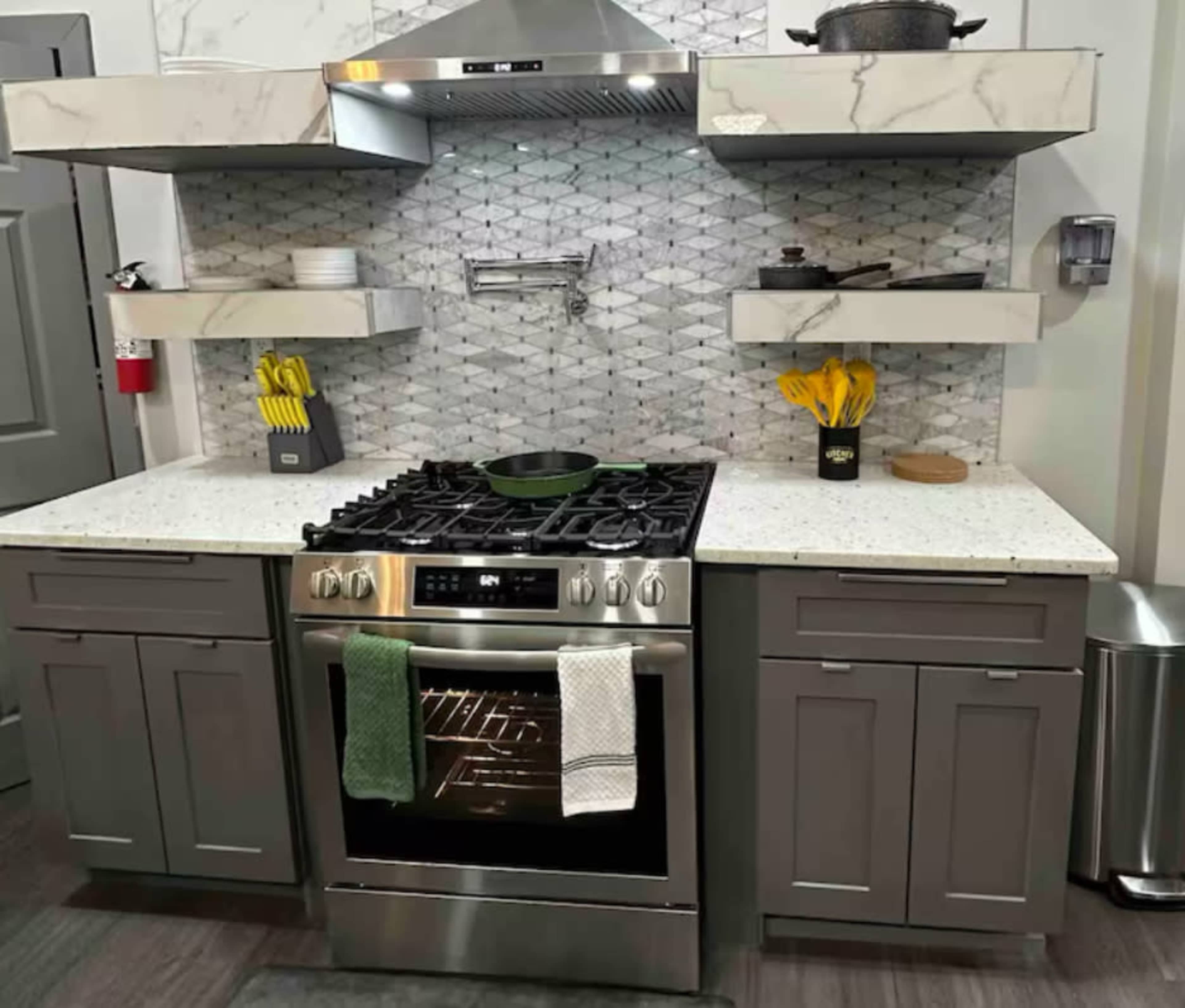 The image shows a modern kitchen with a gas stove, a stainless steel oven, marble backsplash, and gray cabinetry featuring open shelves with cooking utensils and a pot.