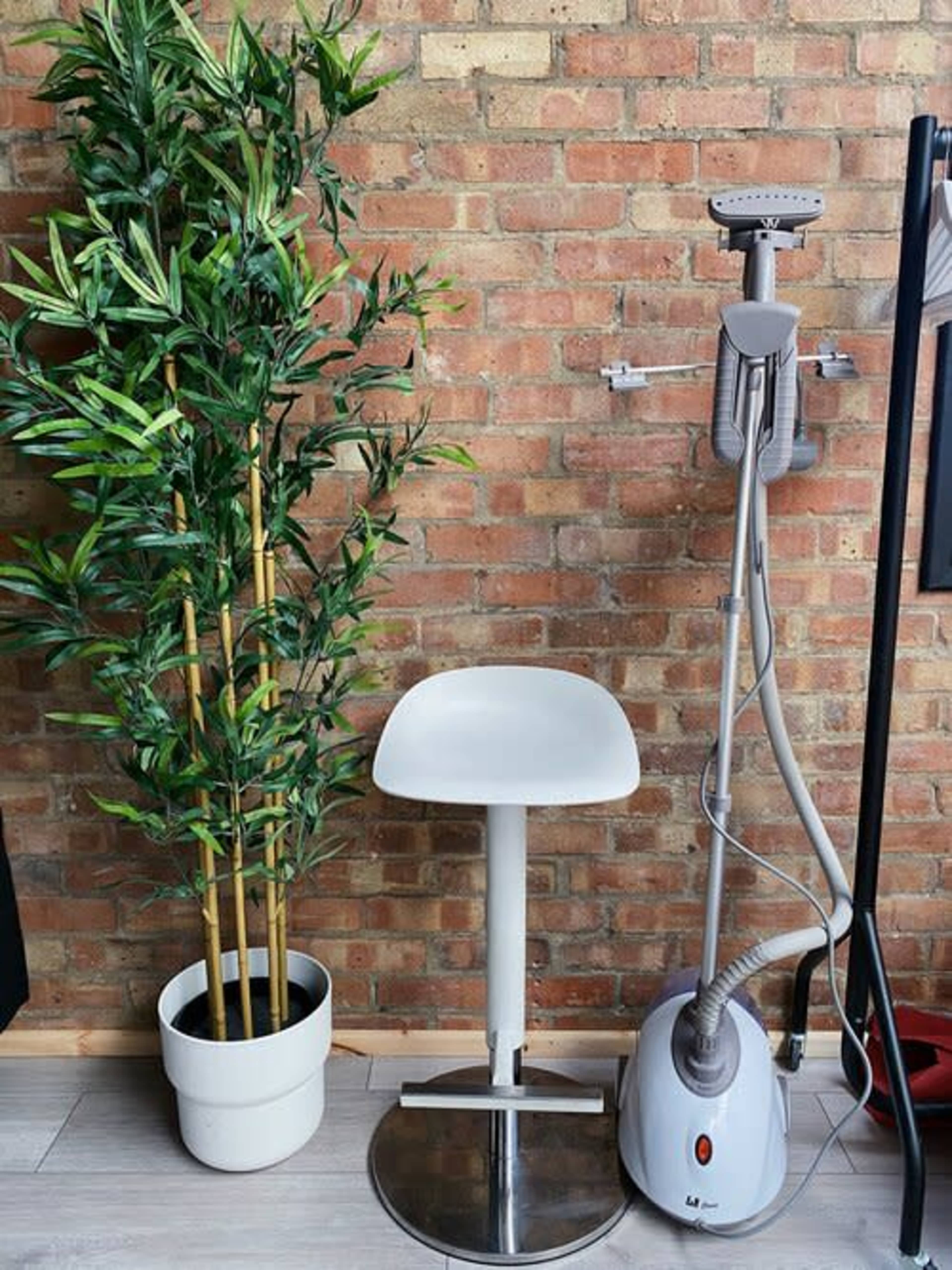 A white stool beside a potted plant and a steam cleaner against a brick wall.