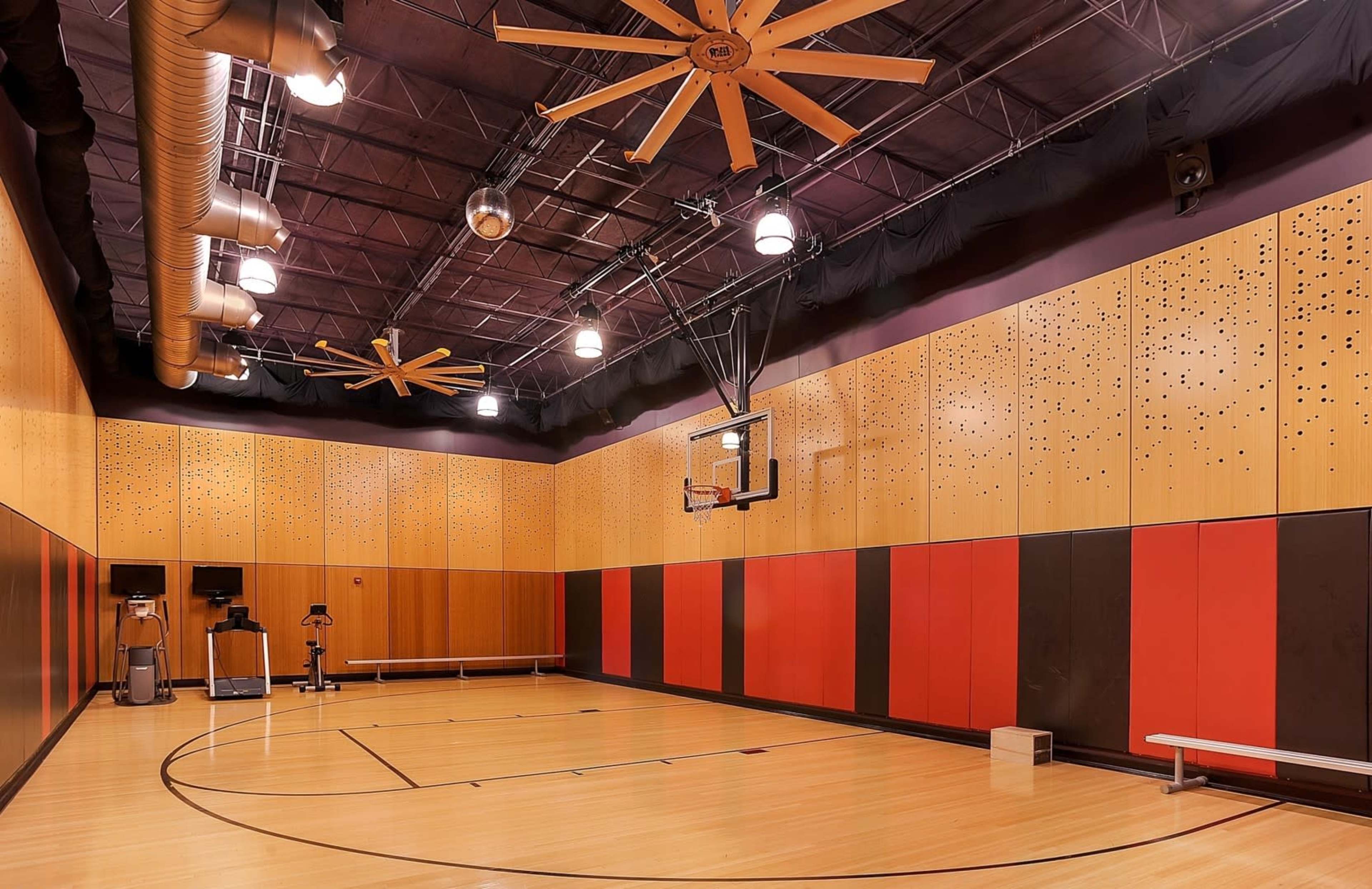 The image shows an indoor basketball court with wooden flooring, large ceiling fans, and walls decorated in a pattern of orange, black, and wood paneling.