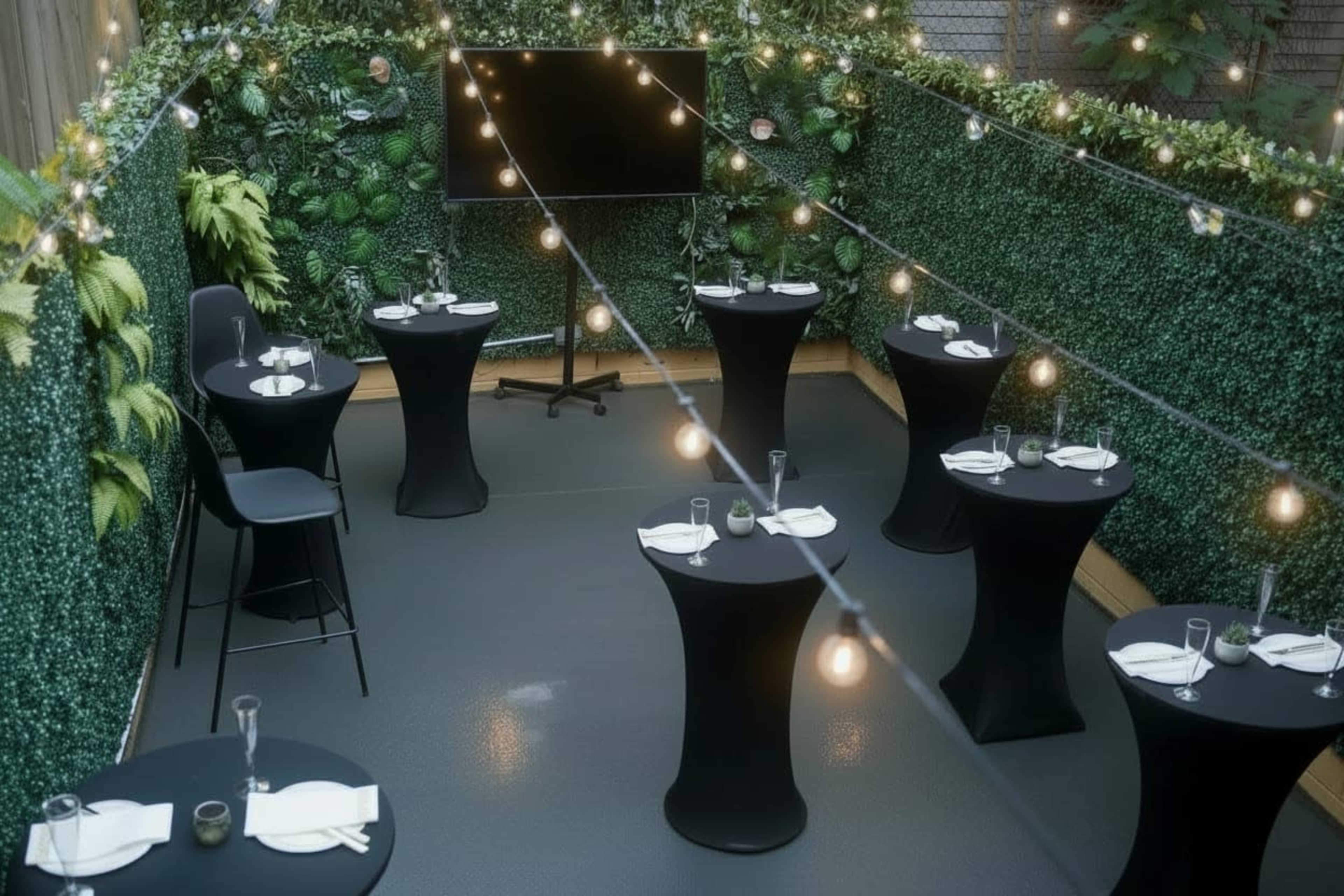 The image shows a decorated outdoor event space featuring several round black tables, a television screen, and string lights above, surrounded by greenery.