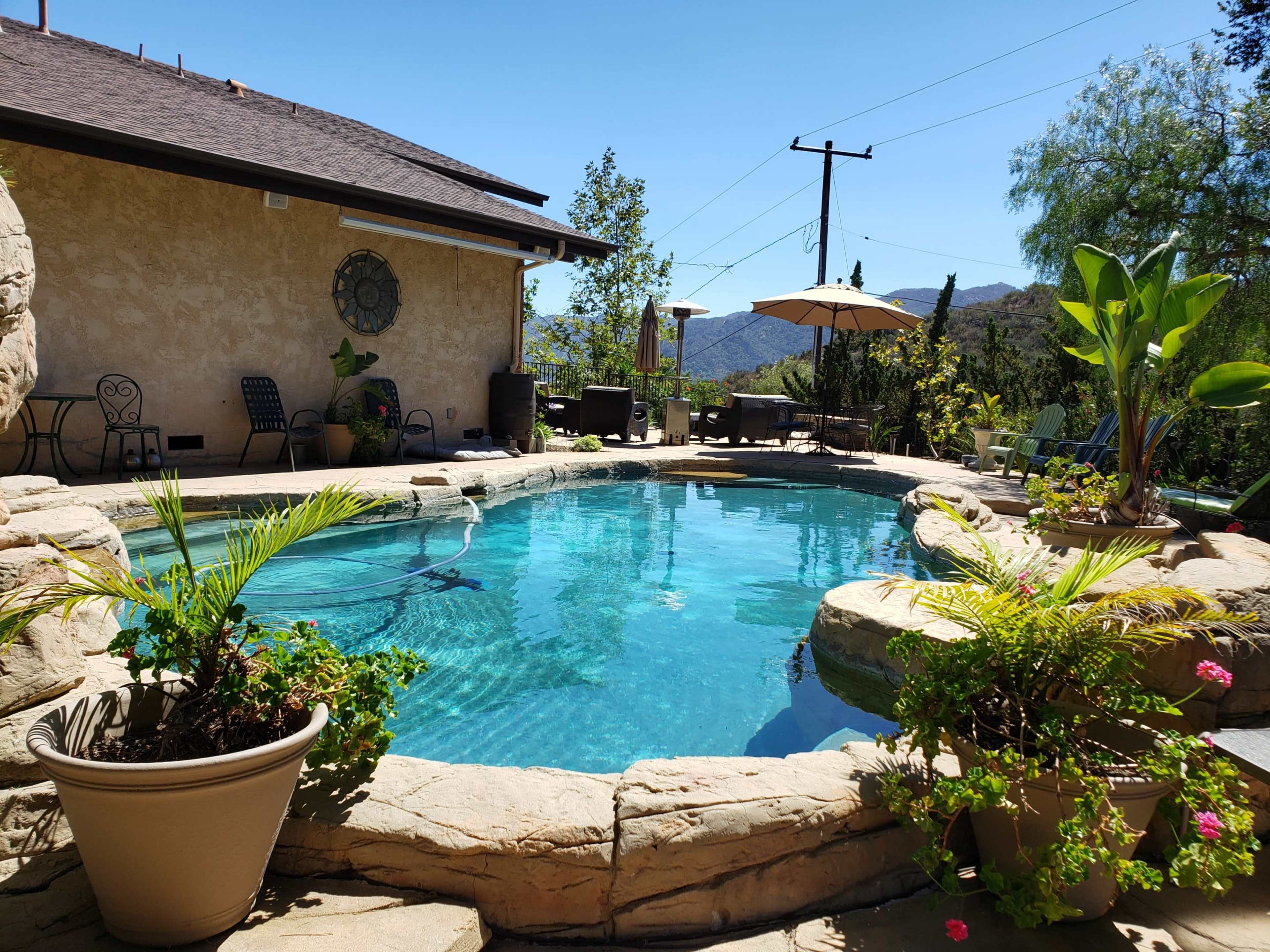 A swimming pool surrounded by rocky landscaping, potted plants, and patio furniture, with mountains visible in the background.