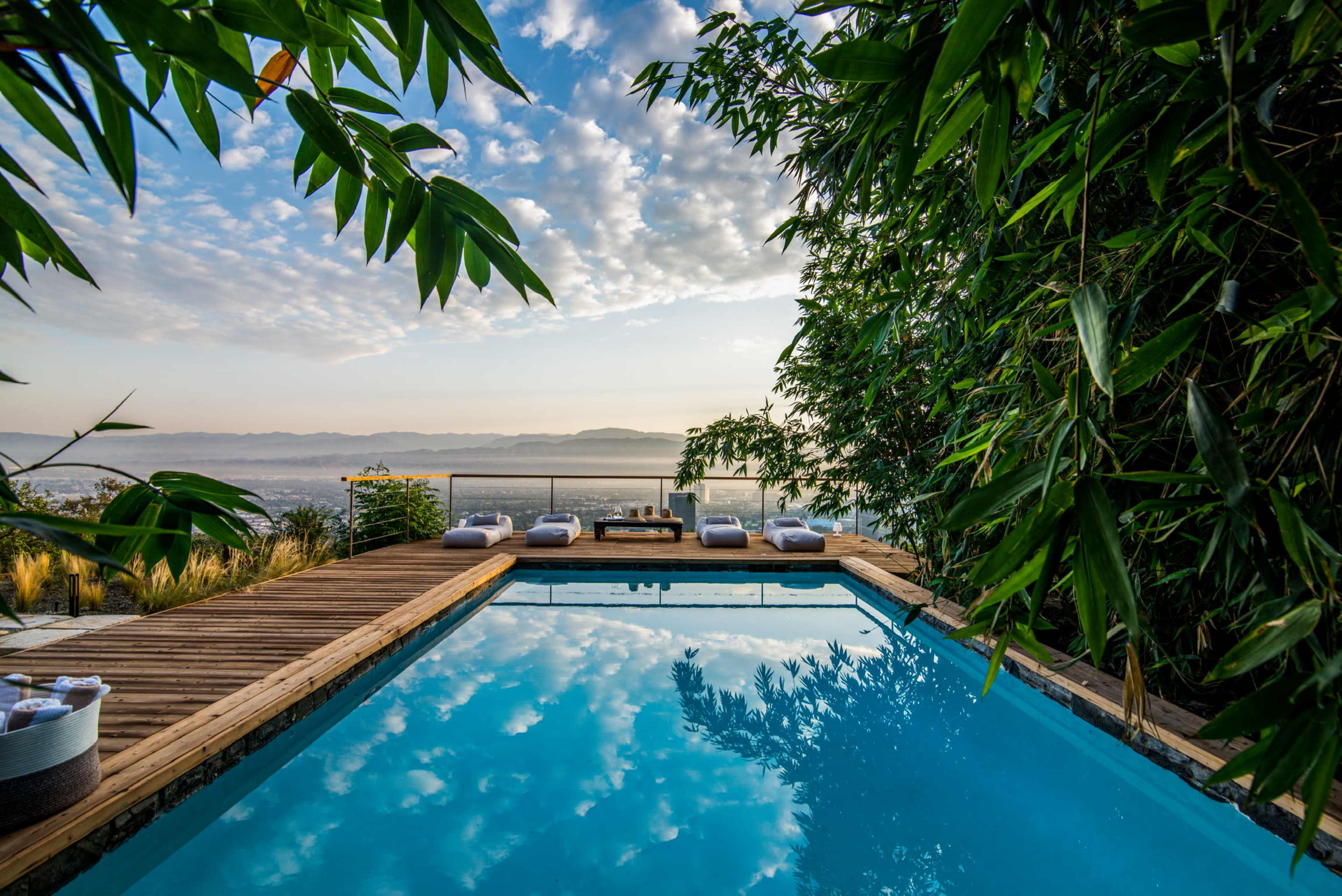 A swimming pool with a wooden deck is surrounded by greenery and offers a view of the mountains under a cloudy sky.