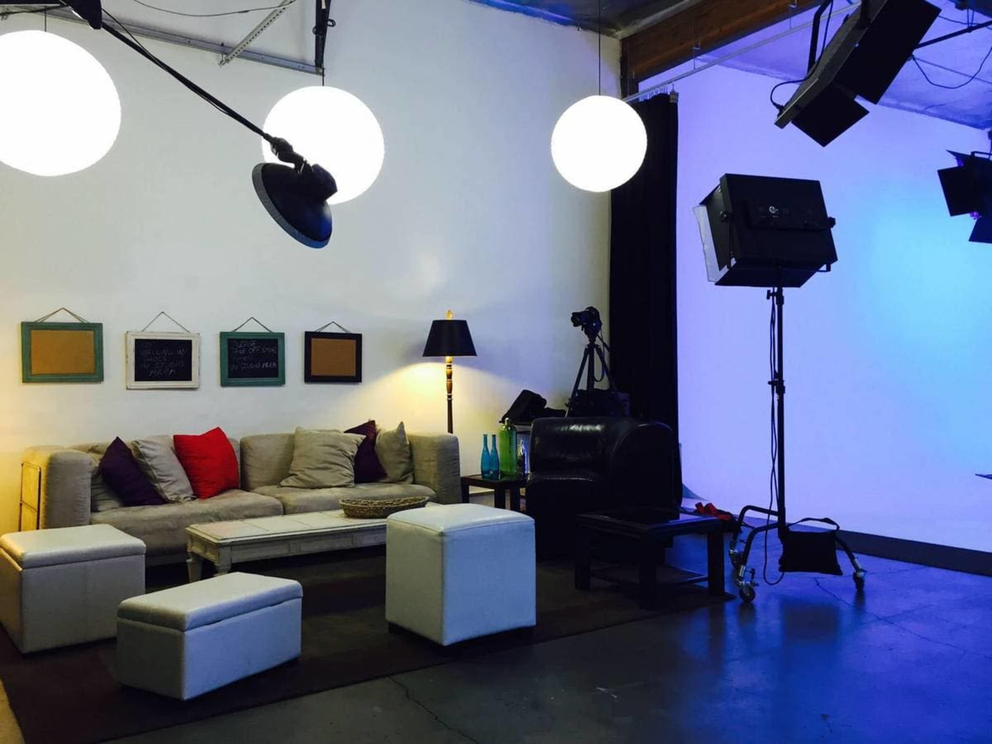A studio setup featuring a beige sofa with colorful pillows, white ottomans, a floor lamp, and various lighting equipment against a plain background.