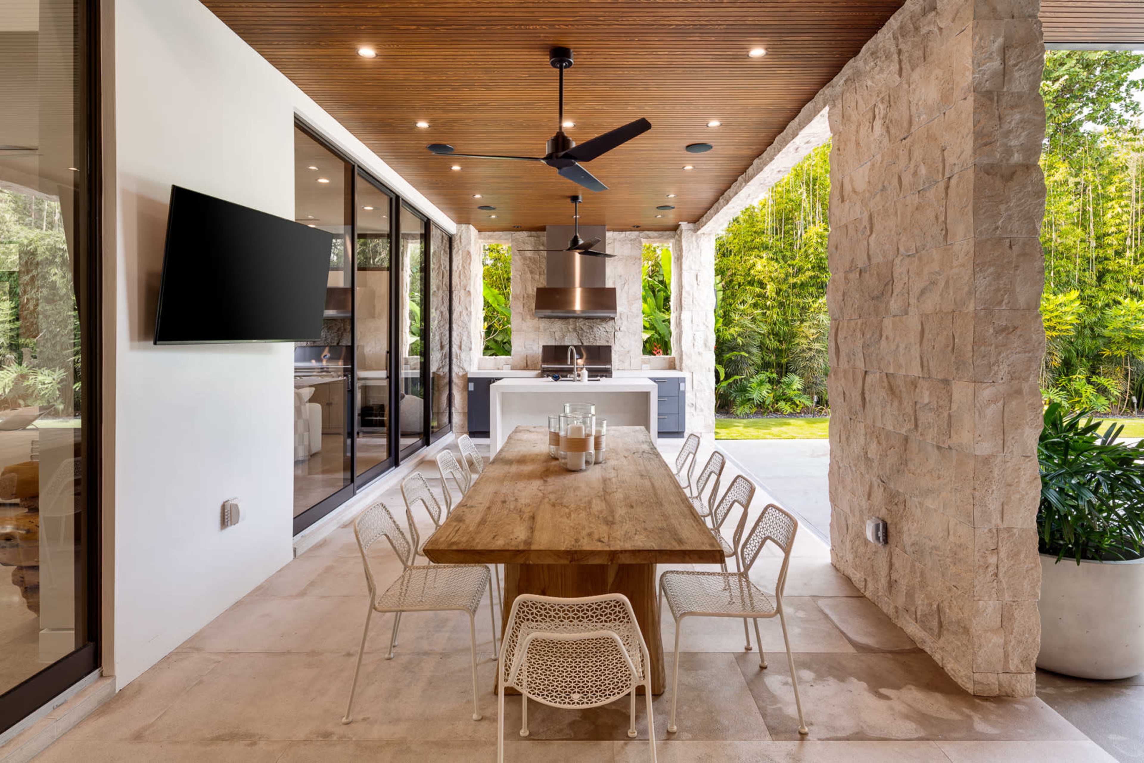 An outdoor dining area features a long wooden table surrounded by white chairs, with a modern kitchen and lush greenery in the background.