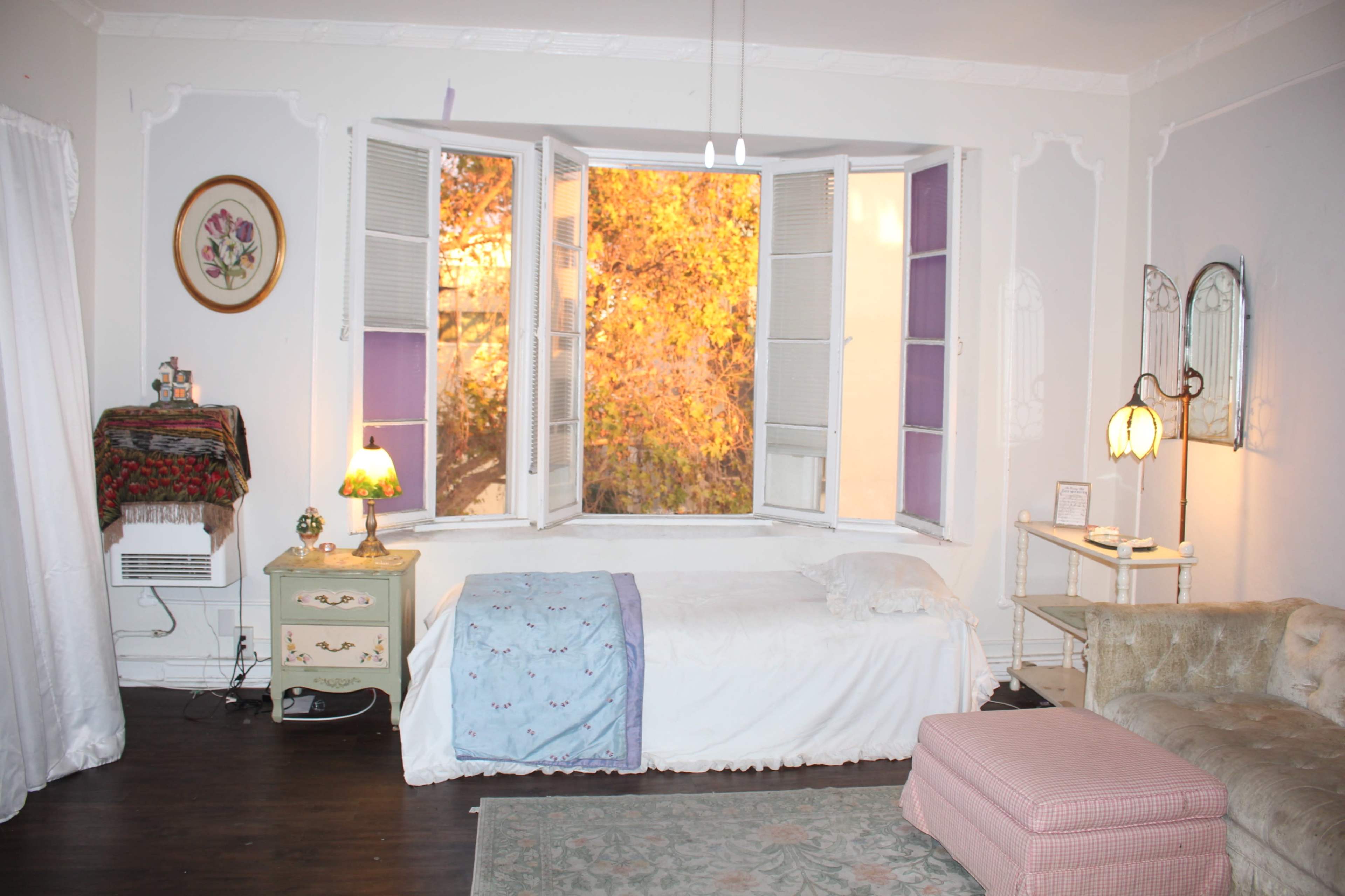 A cozy bedroom features a wooden floor and large windows with purple shutters, showcasing a view of autumn foliage outside.