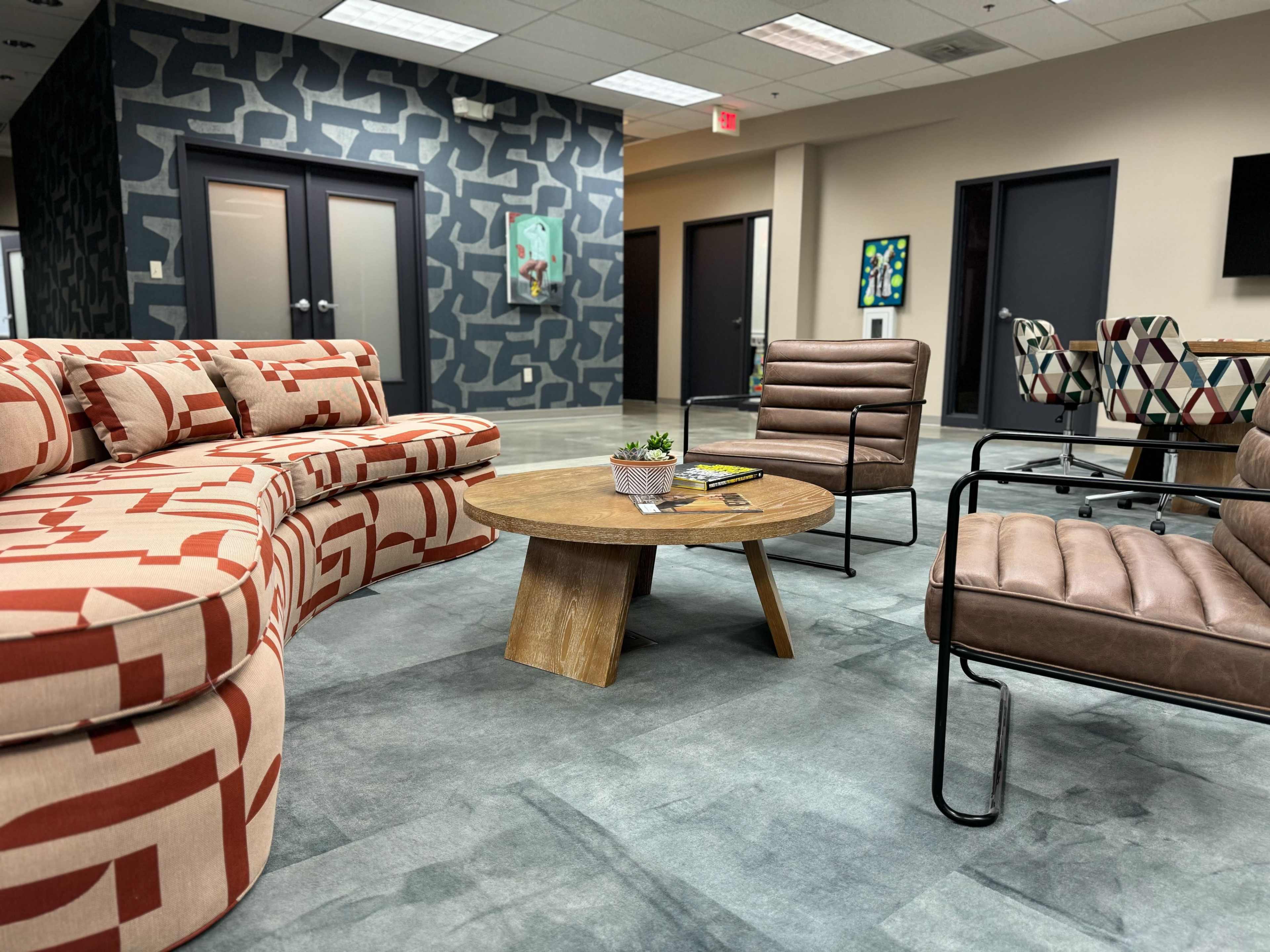 A modern lounge area featuring a patterned sectional sofa, a wooden coffee table, and leather chairs, set against a backdrop of geometric wallpaper.