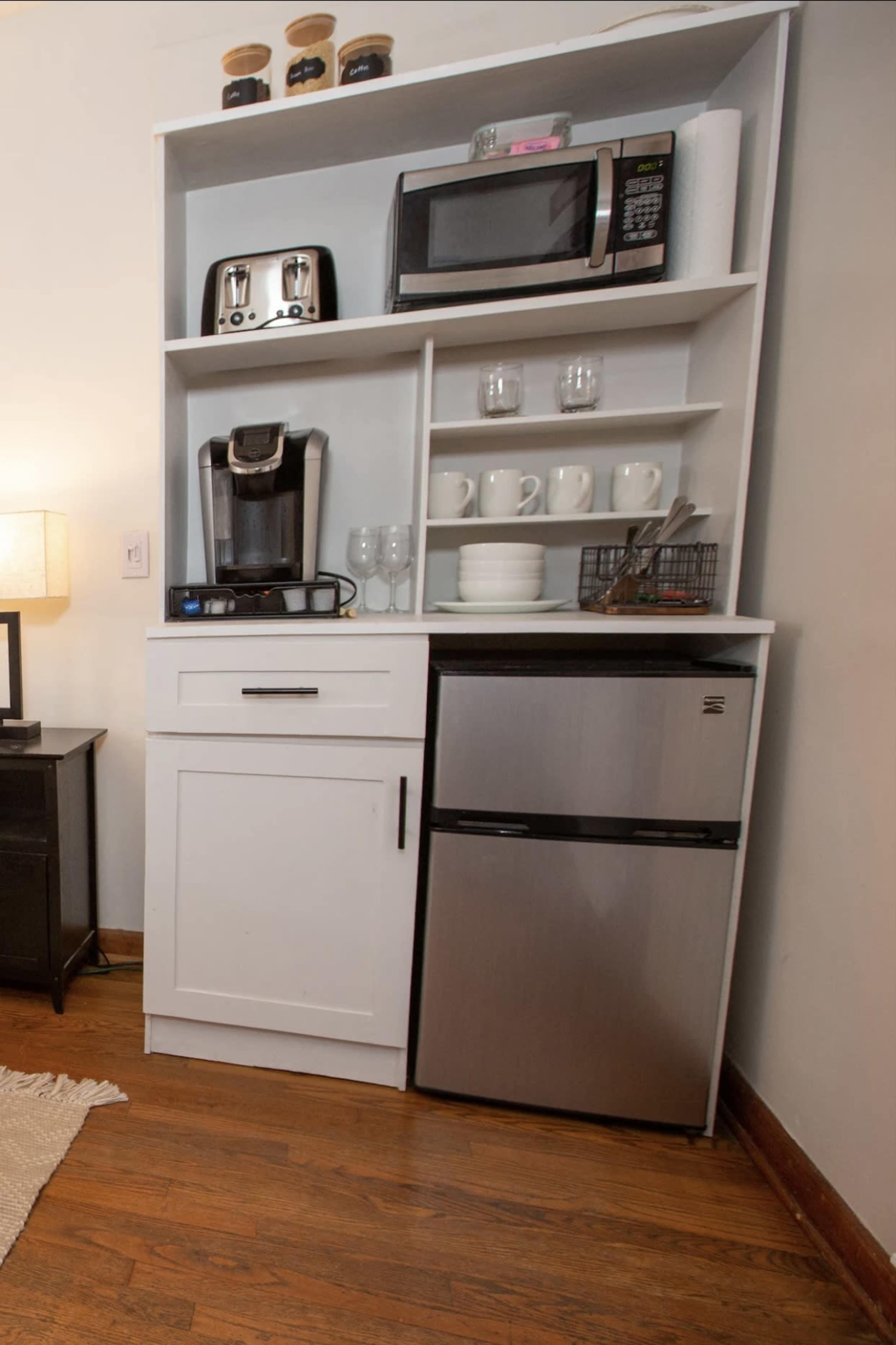 The image shows a white kitchenette unit with a coffee maker, microwave, and a small refrigerator, along with shelves holding mugs and glasses.
