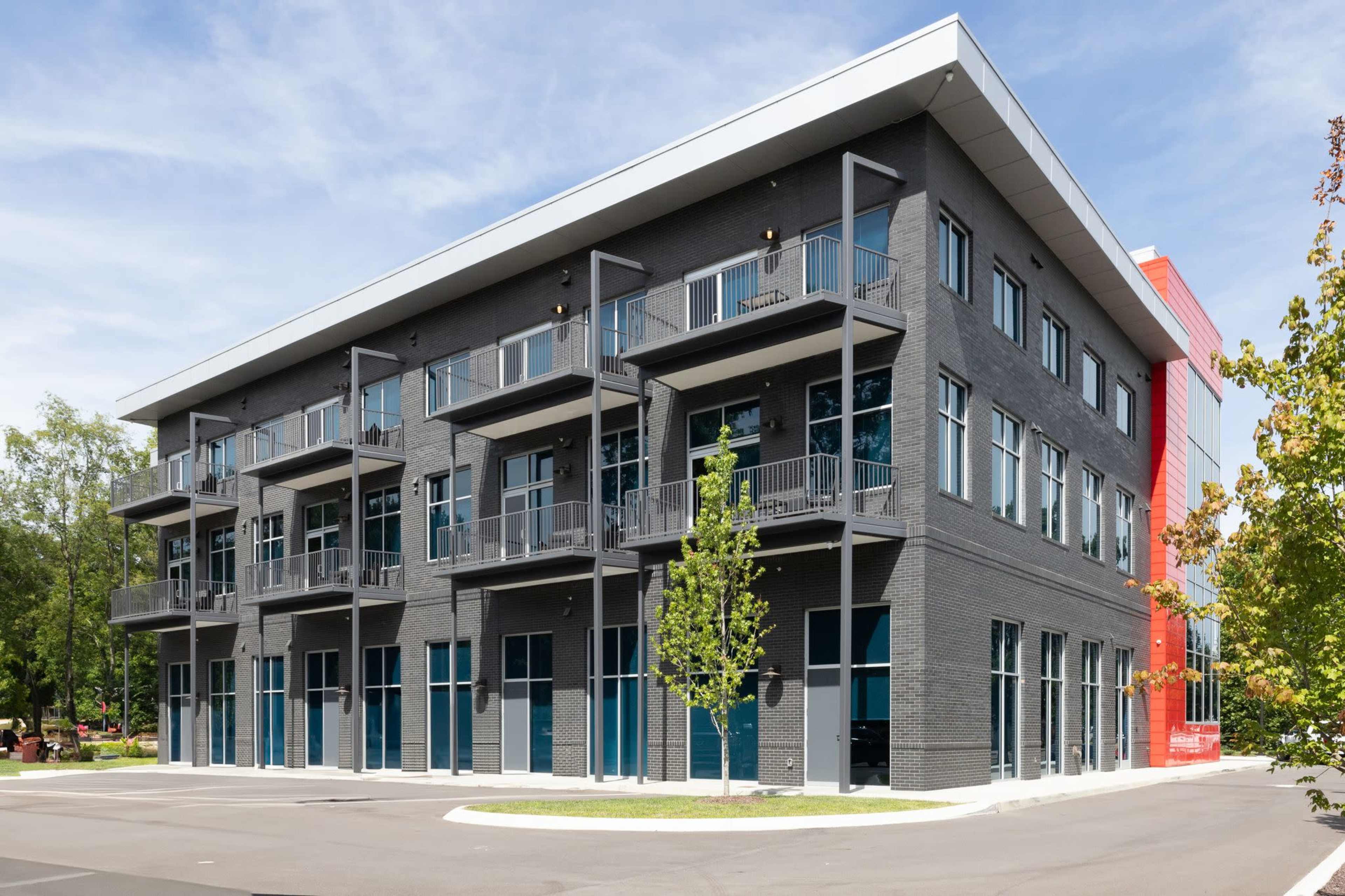 A modern three-story commercial building with a grey brick facade, balconies, large windows, and a section painted in red.