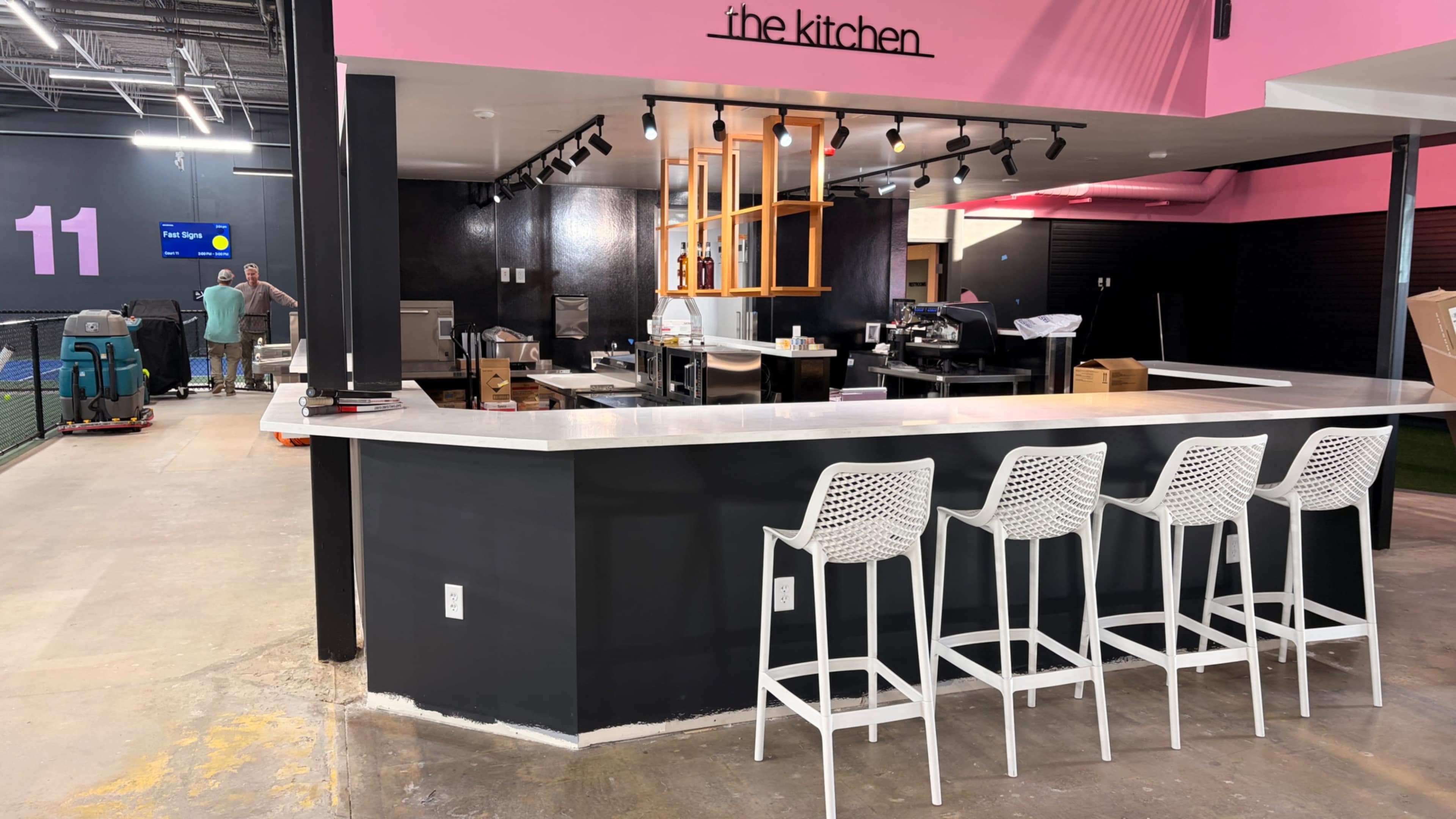The image shows a modern kitchen and dining area with a countertop bar featuring white stools, vibrant pink walls, and a well-equipped cooking space in the background.