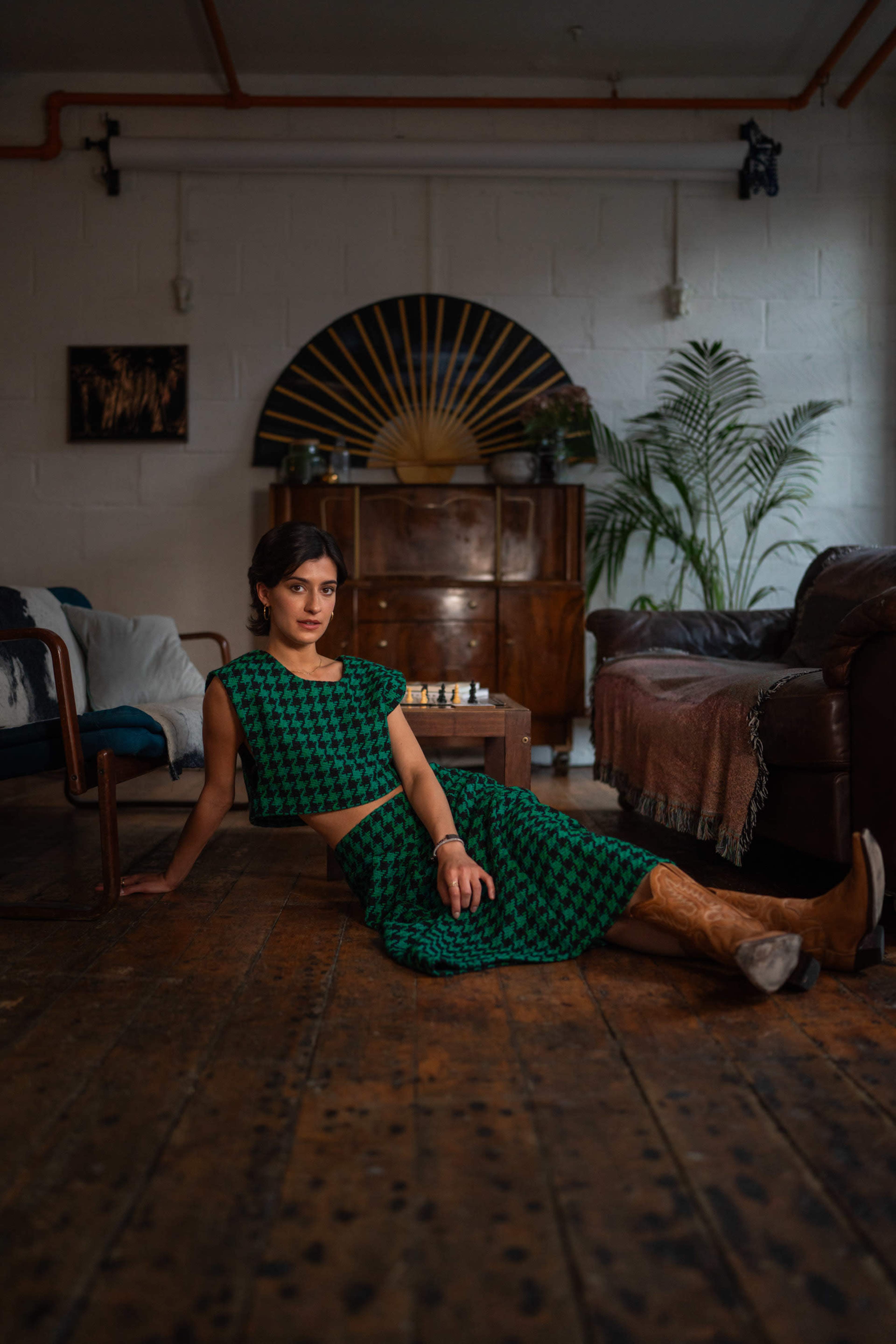 A woman wearing a green cropped outfit sits on a wooden floor in a room with vintage furnishings and plants.