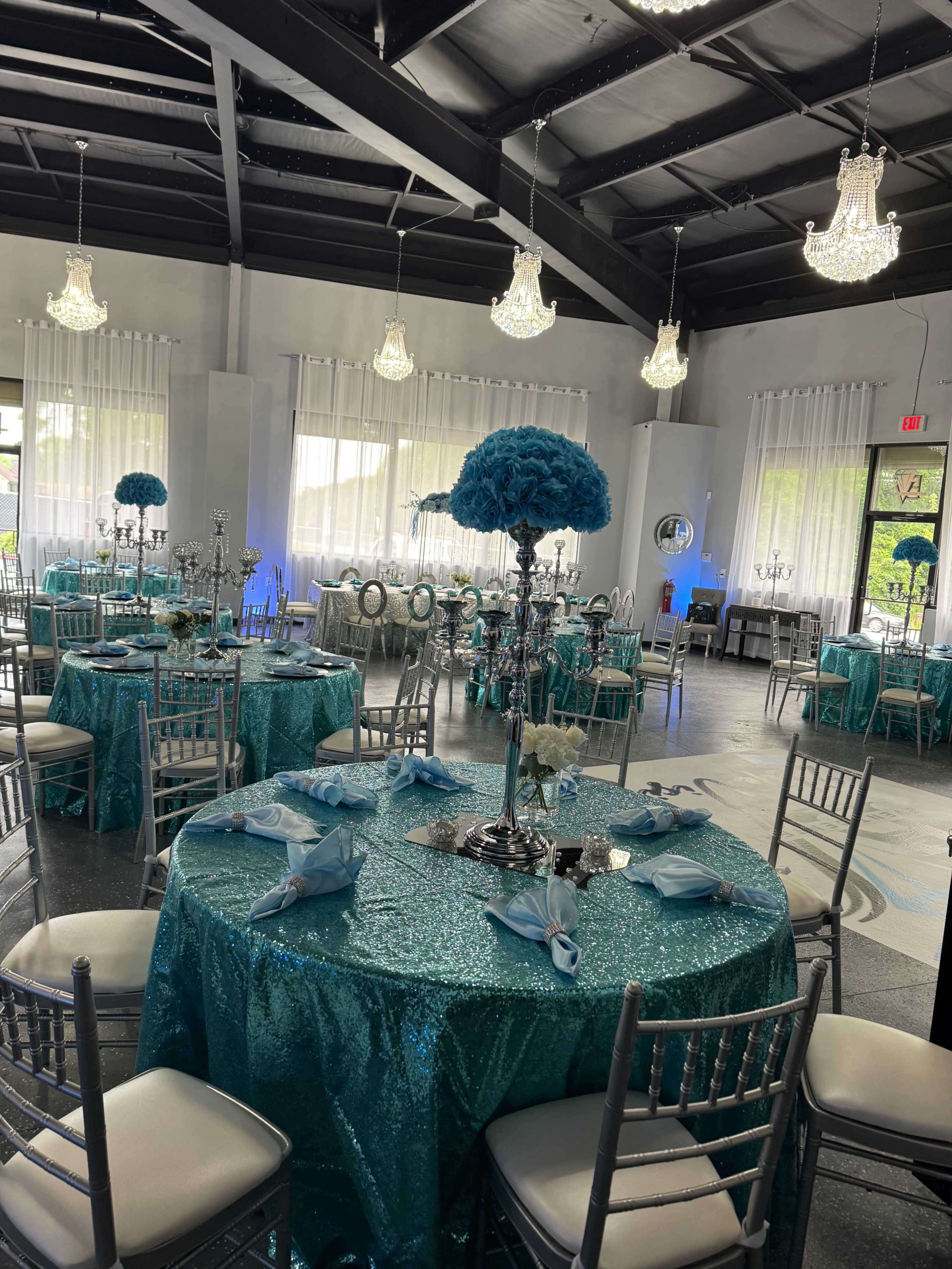 A banquet hall is set up with teal sequin tablecloths, silver candelabras, and tall floral centerpieces on each table under sparkling chandeliers.