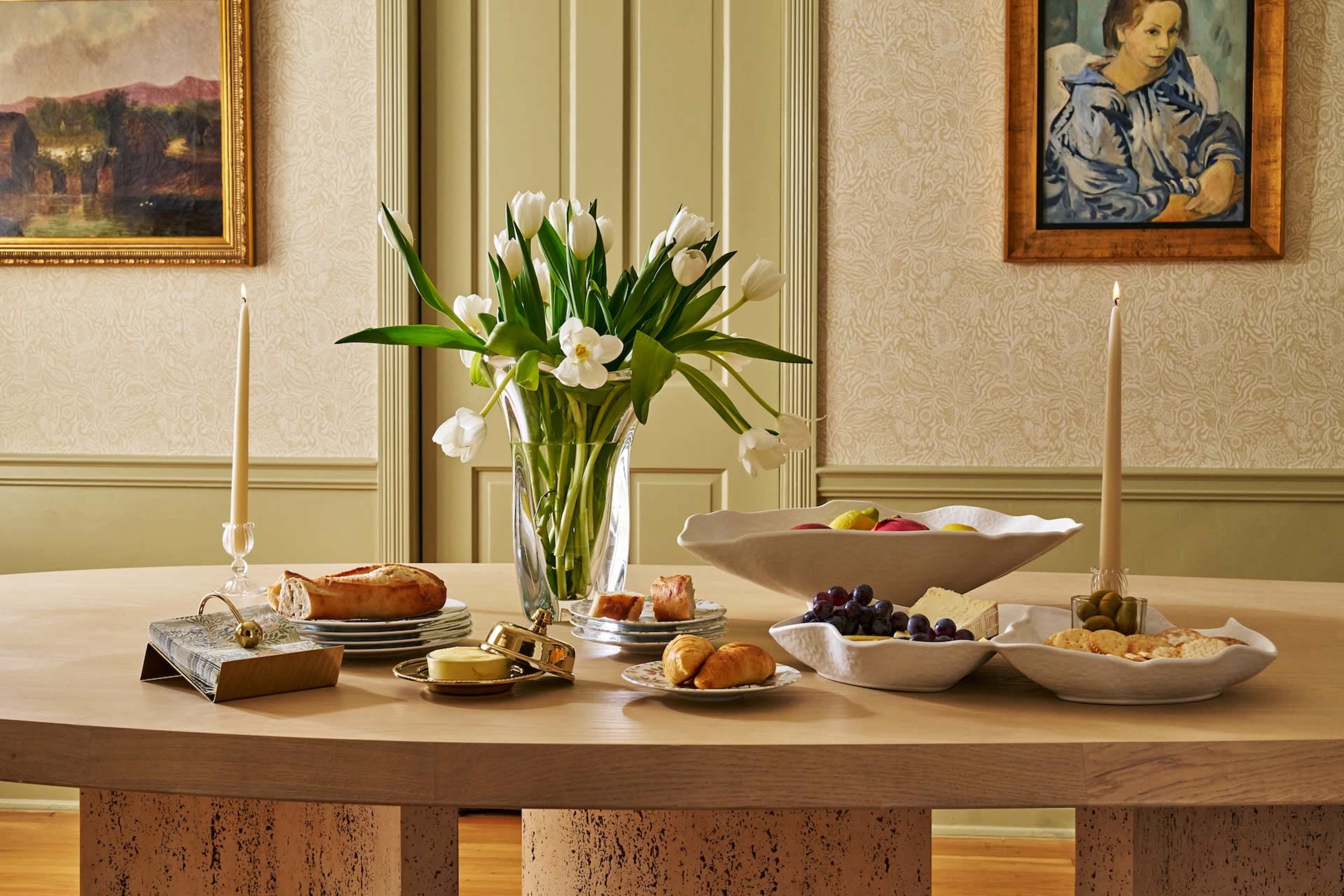 A round wooden table is set with an array of food items, white flowers in a vase, and candles, against a backdrop of patterned wallpaper and artwork.