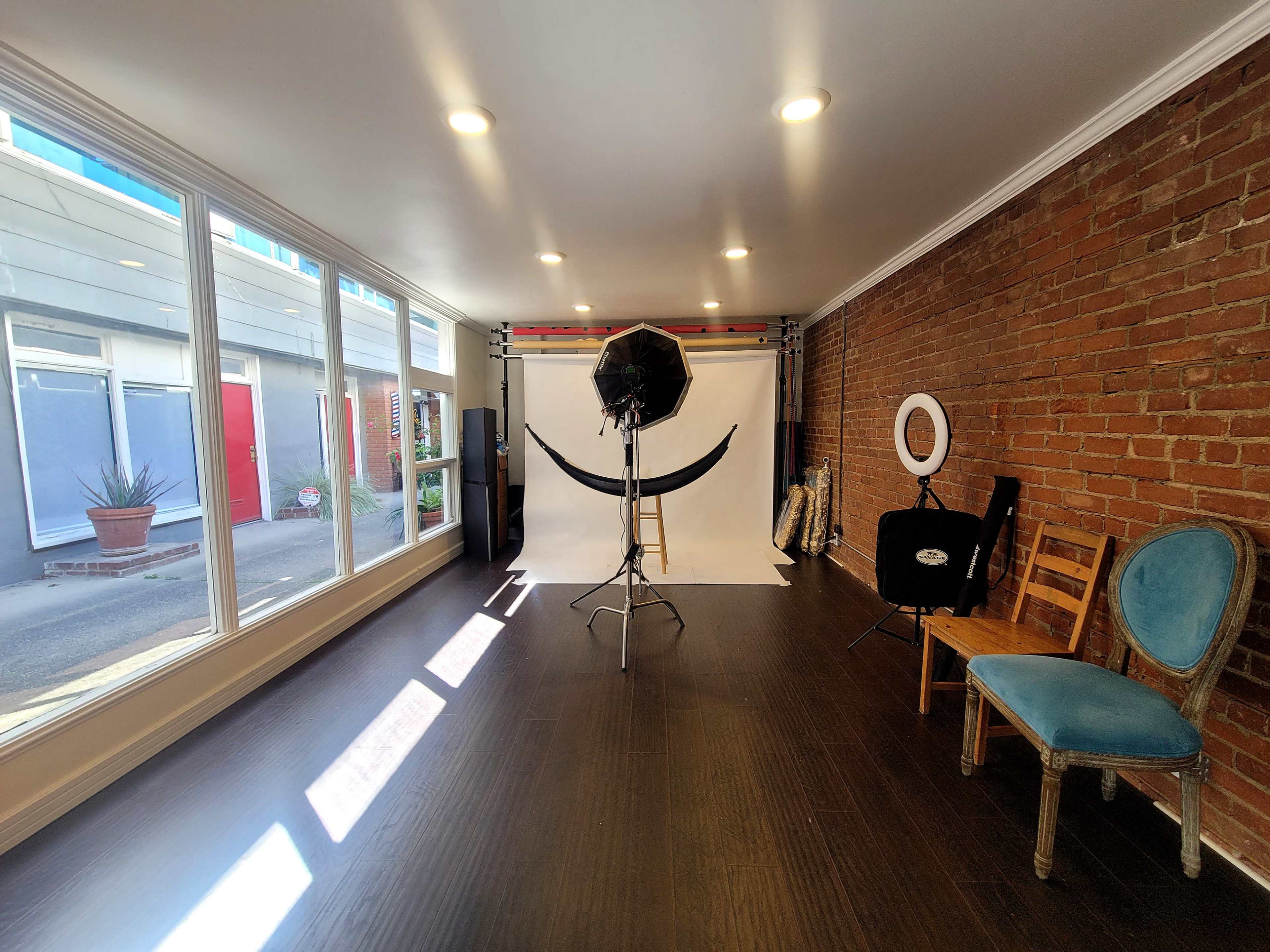 The image shows a spacious photography studio featuring a white backdrop, lighting equipment, and a mix of chairs against a brick wall, with natural light coming in through large windows.